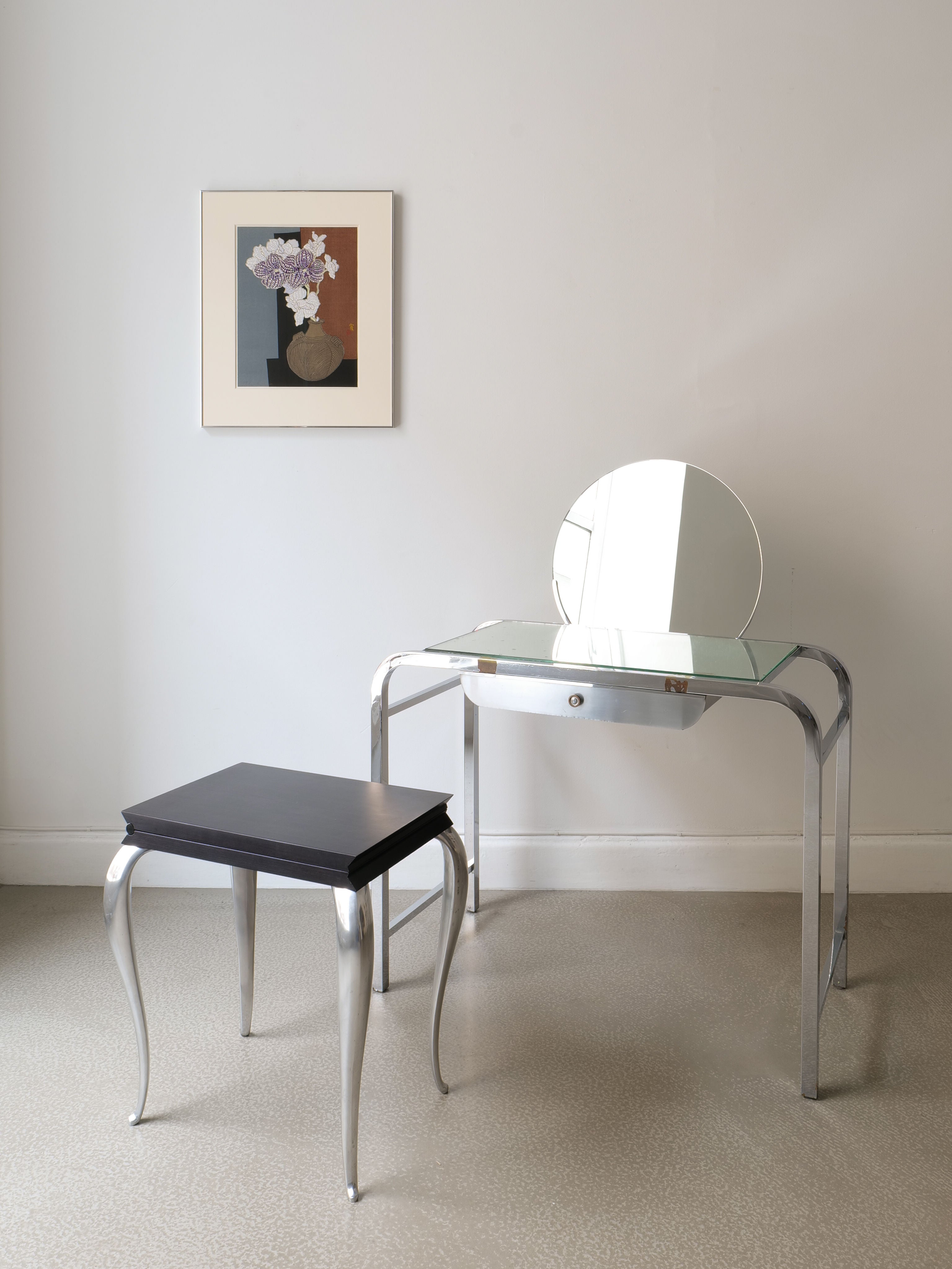 A modern dressing table with a round silver-framed mirror is set against a light gray wall. A matching stool with curved legs sits gracefully in front, while above, the Woodcut Print by Tokuriki Tomikichiro from Collection apart adds an artful charm.