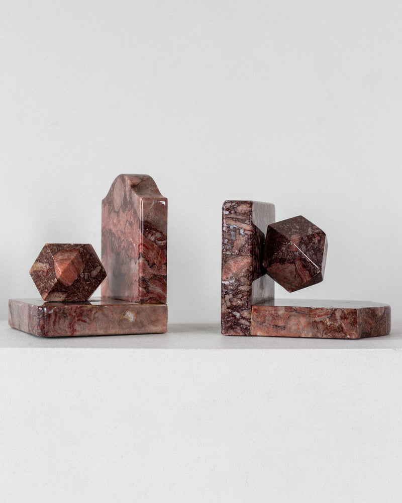 The Pair of Art Deco Marble Bookends by Spigel features two pieces made from marbled brown and red stone. Each bookend has a rectangular base with a unique 3D shape, reminiscent of abstract geometric sculptures, displayed on a plain white surface.