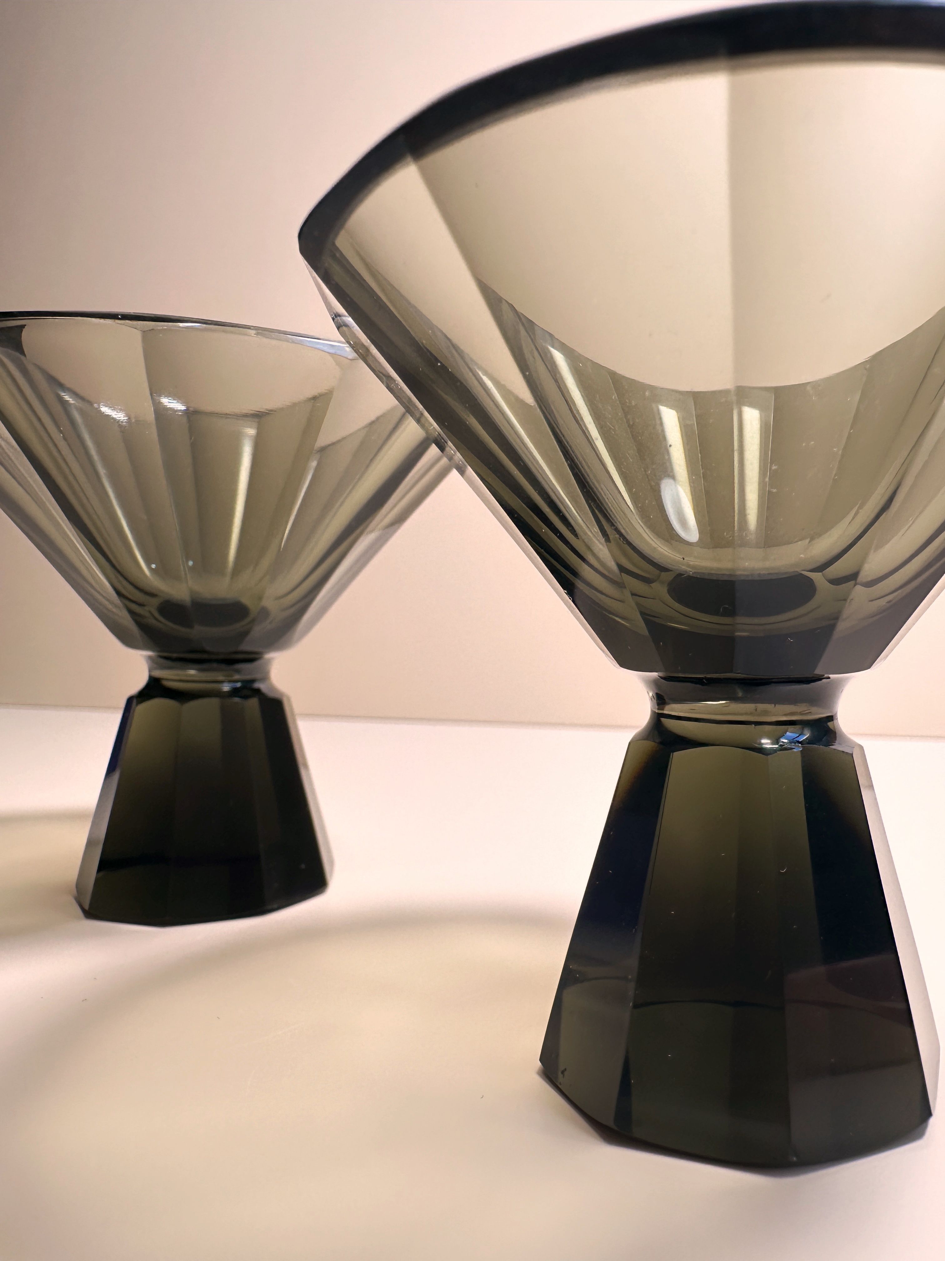 A close-up image of two elegant, smokey-gray 1930s Art Deco Facetted Liqueur Glasses by Dodo Vintage, featuring wide, funnel-shaped bowls and intricately faceted pedestal bases. These handmade goblets are positioned diagonally, providing a striking view of their detailed design. The background is a soft, off-white color.