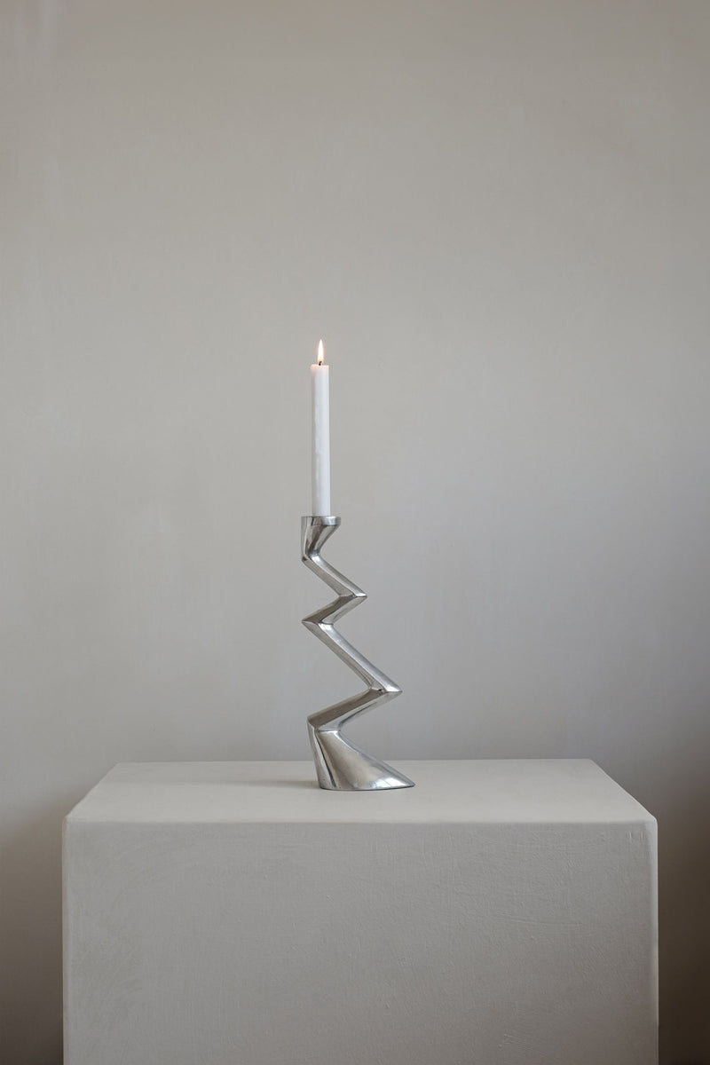 A single white candle glows in the Matthew Hilton ZigZag Candle Holder Large by septembre studios, its angular silhouette atop a light pedestal against a neutral wall—an ideal accent for contemporary interiors.