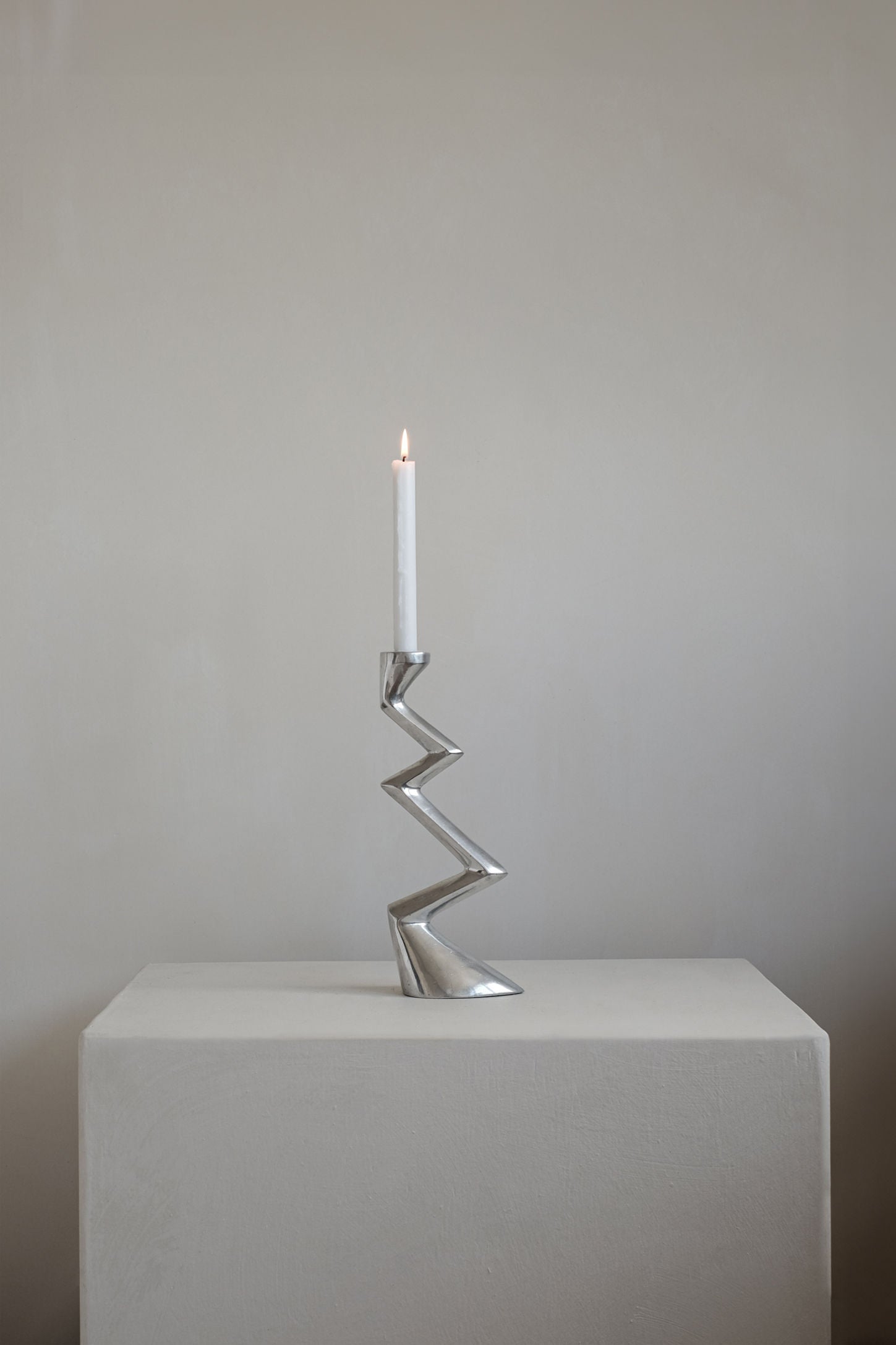 A single white candle glows in the Matthew Hilton ZigZag Candle Holder Large by septembre studios, its angular silhouette atop a light pedestal against a neutral wall—an ideal accent for contemporary interiors.