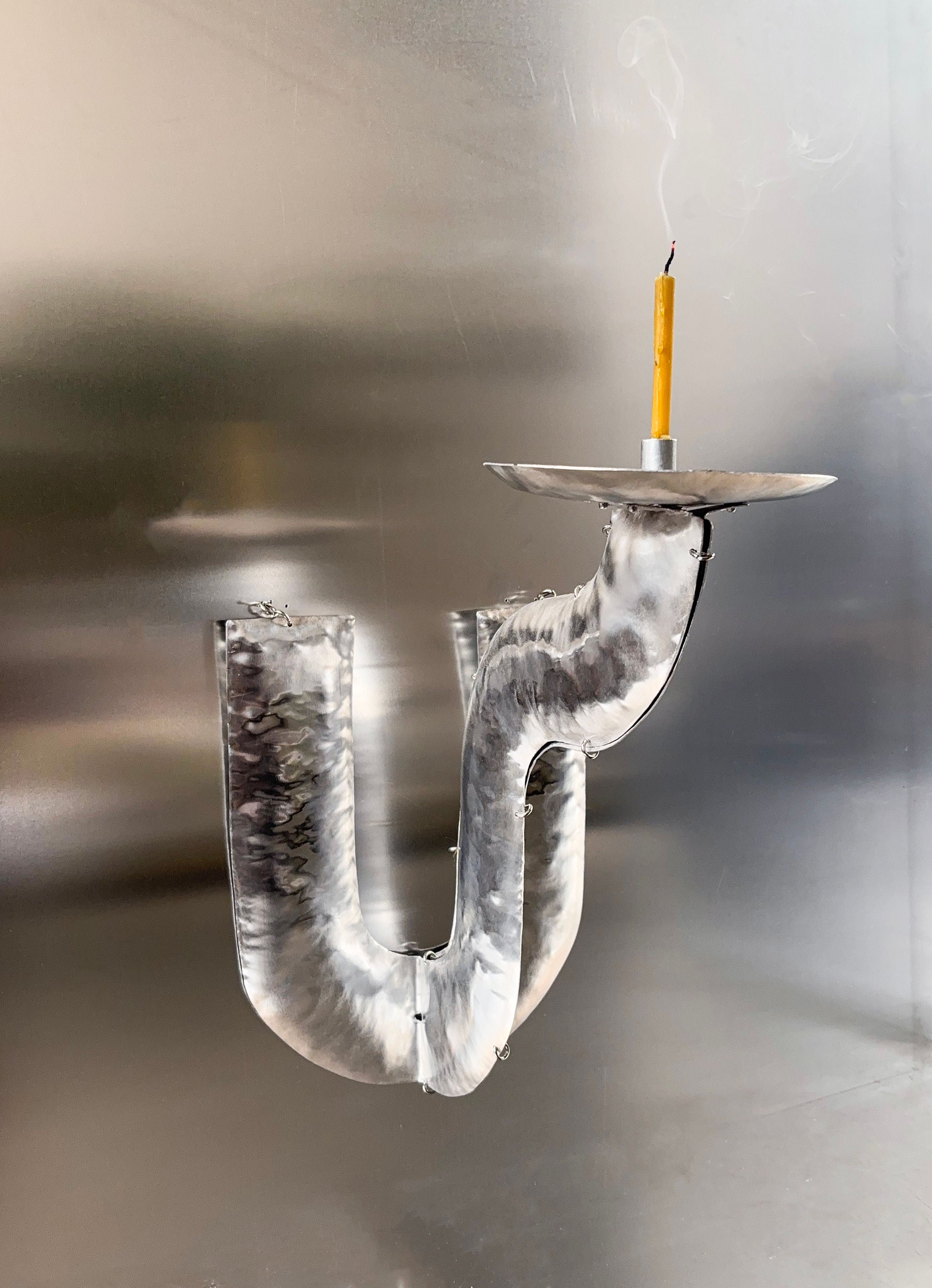 The Phaedra candle holder WALL by Bregje Sliepenbeek features a candle on a metallic J-shaped pipe with its smoke rising against a sleek, reflective aluminum background. Handmade in Amsterdam, it brings artisanal elegance to any space.