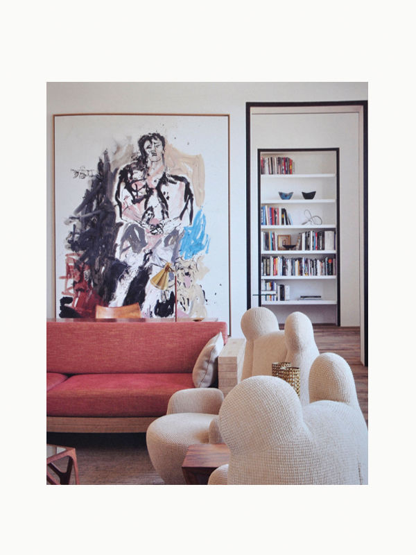 A cozy living room featuring a red sofa, a unique Maison Plage armchair designed by Pierre Yovanovitch, and an abstract painting on the left wall. On the right, a white bookshelf filled with books.