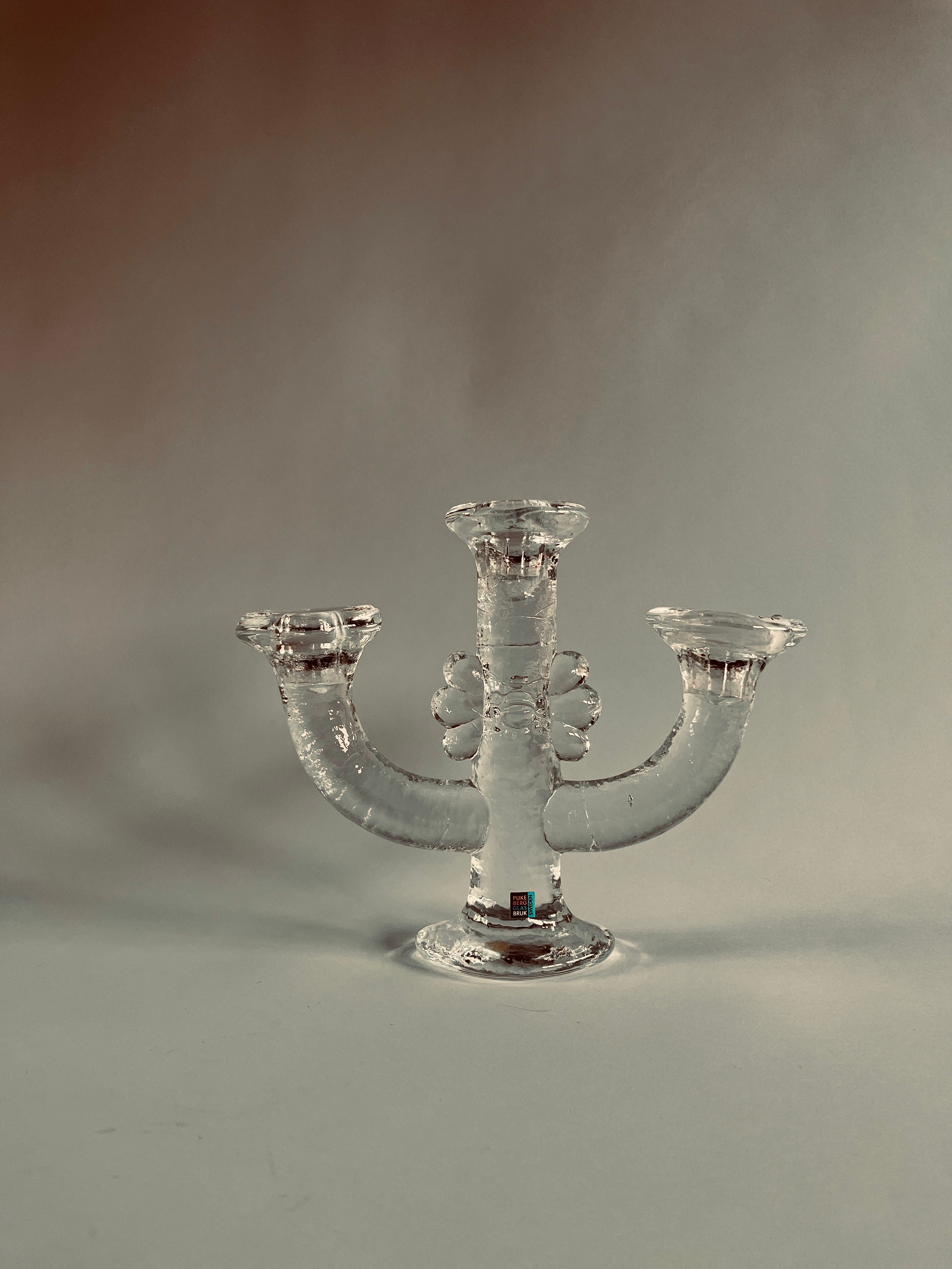 The Maud Vaughan Swedish Glass Candelabra, designed by Staffan Gellerstedt, is set against a neutral background. It features three curved arms and ornate detailing, showcasing a simple yet elegant design.