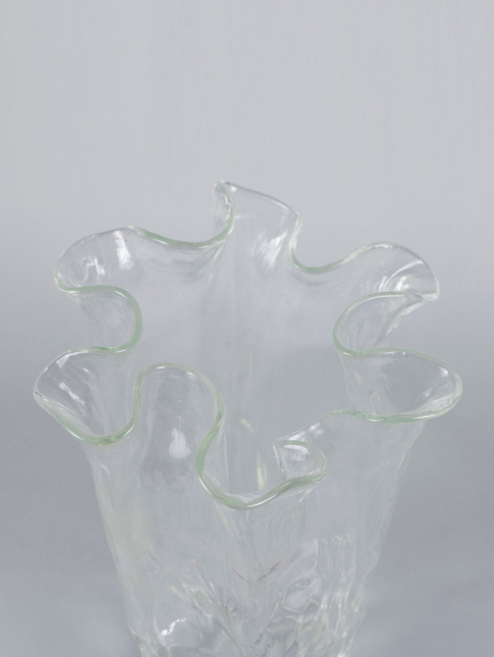 A close-up of the Organic Glass Vase by Pertti Kallioinen from RELIC LONDON showcases its translucent glass with a wavy, ruffled rim and an irregular shape resembling flowing water, creating an organic and fluid appearance against a neutral background.