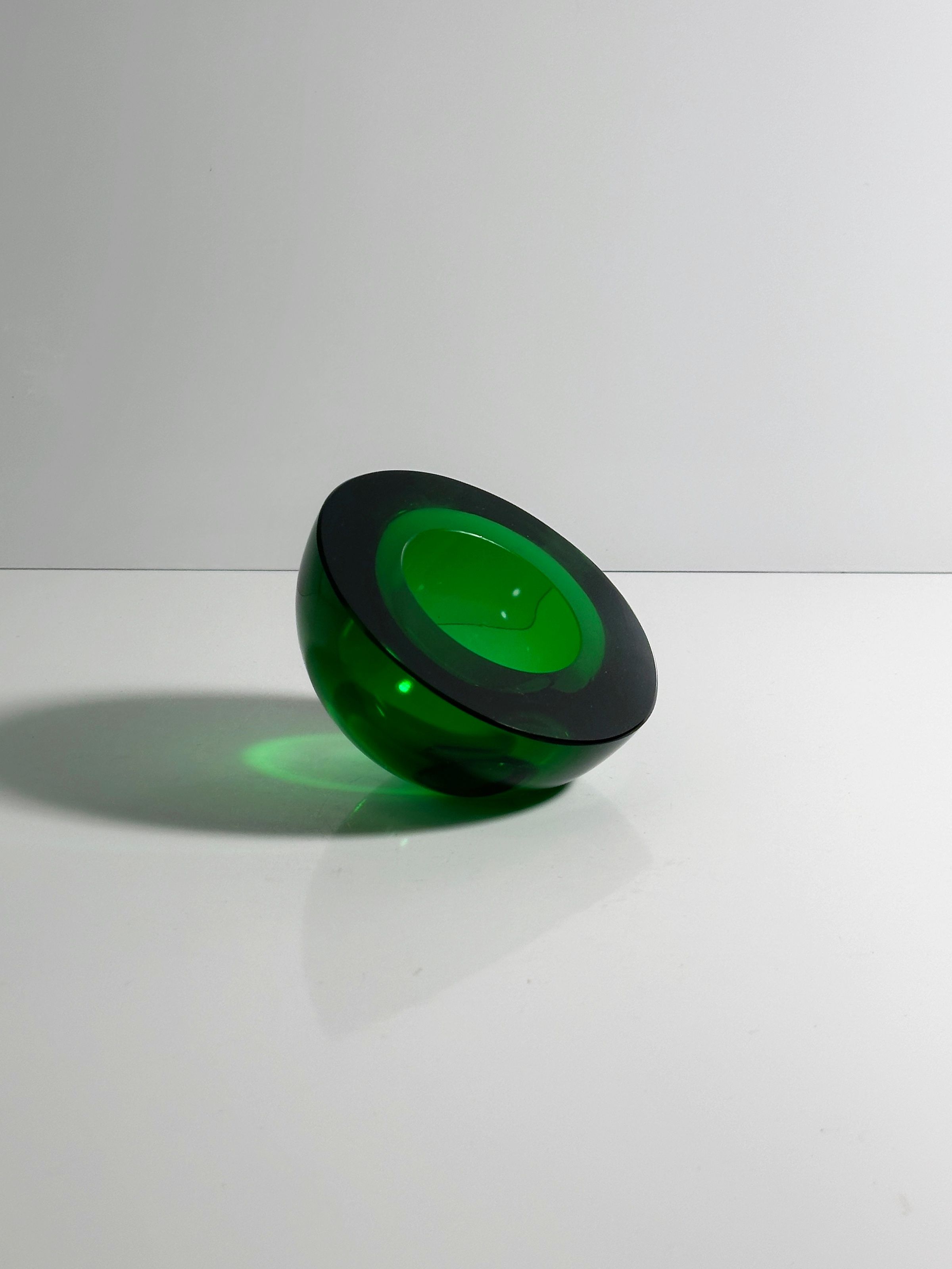 A circular, green glass sculpture by Dodo Vintage, reminiscent of 1960s craftsmanship, sits on a reflective surface. The lighting enhances the transparency and vibrant green hue of this "1960s Space Age Glass Ashtray," casting a slight shadow against a plain background.