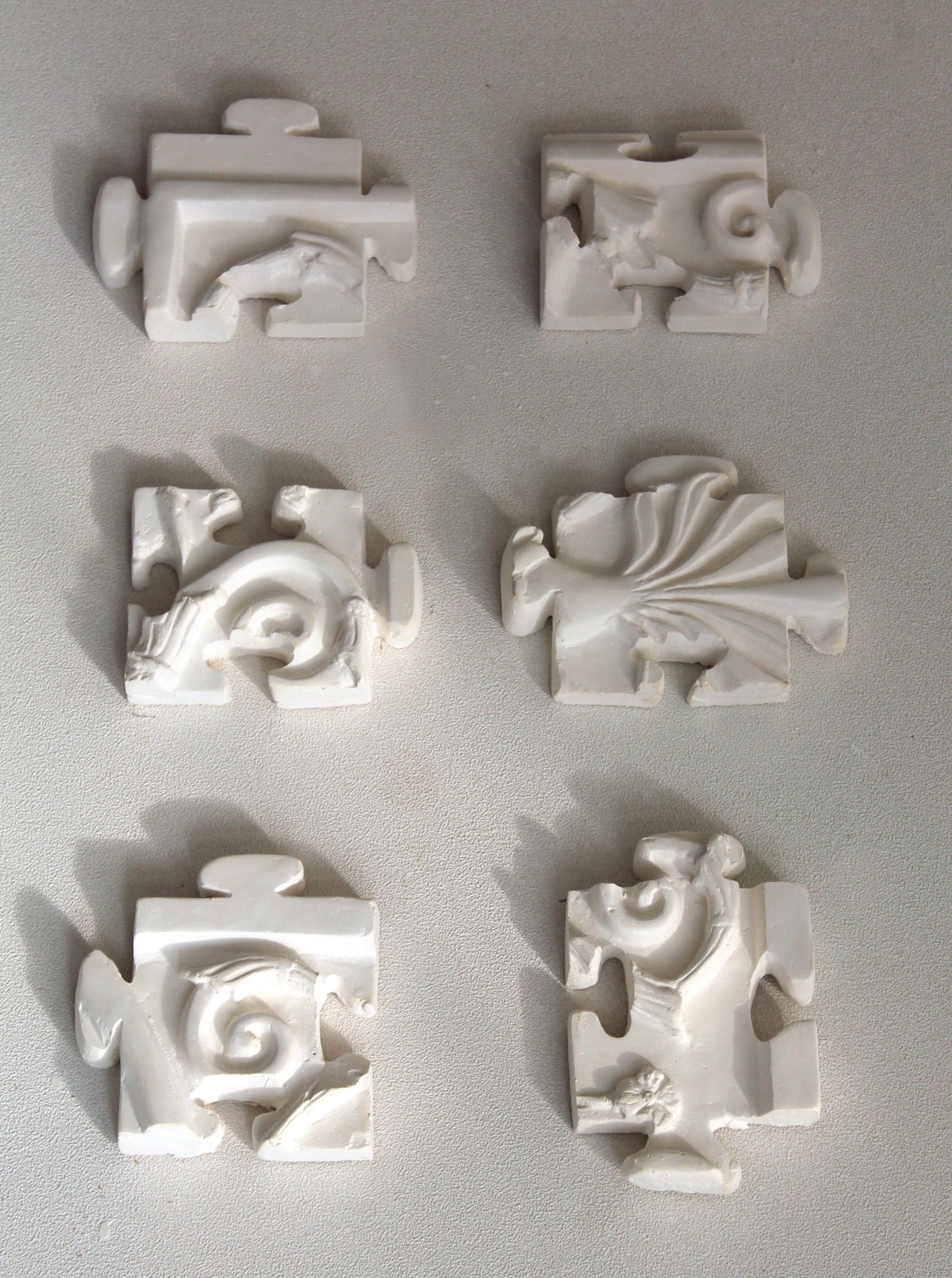 Six small, white plaster relief sculptures from the "Puzzle Antique" collection by Marina Mankarios are arranged in a Sexiptyque, featuring two rows of three. Each piece showcases unique patterns and shapes with abstract and organic forms. They appear to be made of plaster or a similar material and are mounted on a flat, light-colored surface.