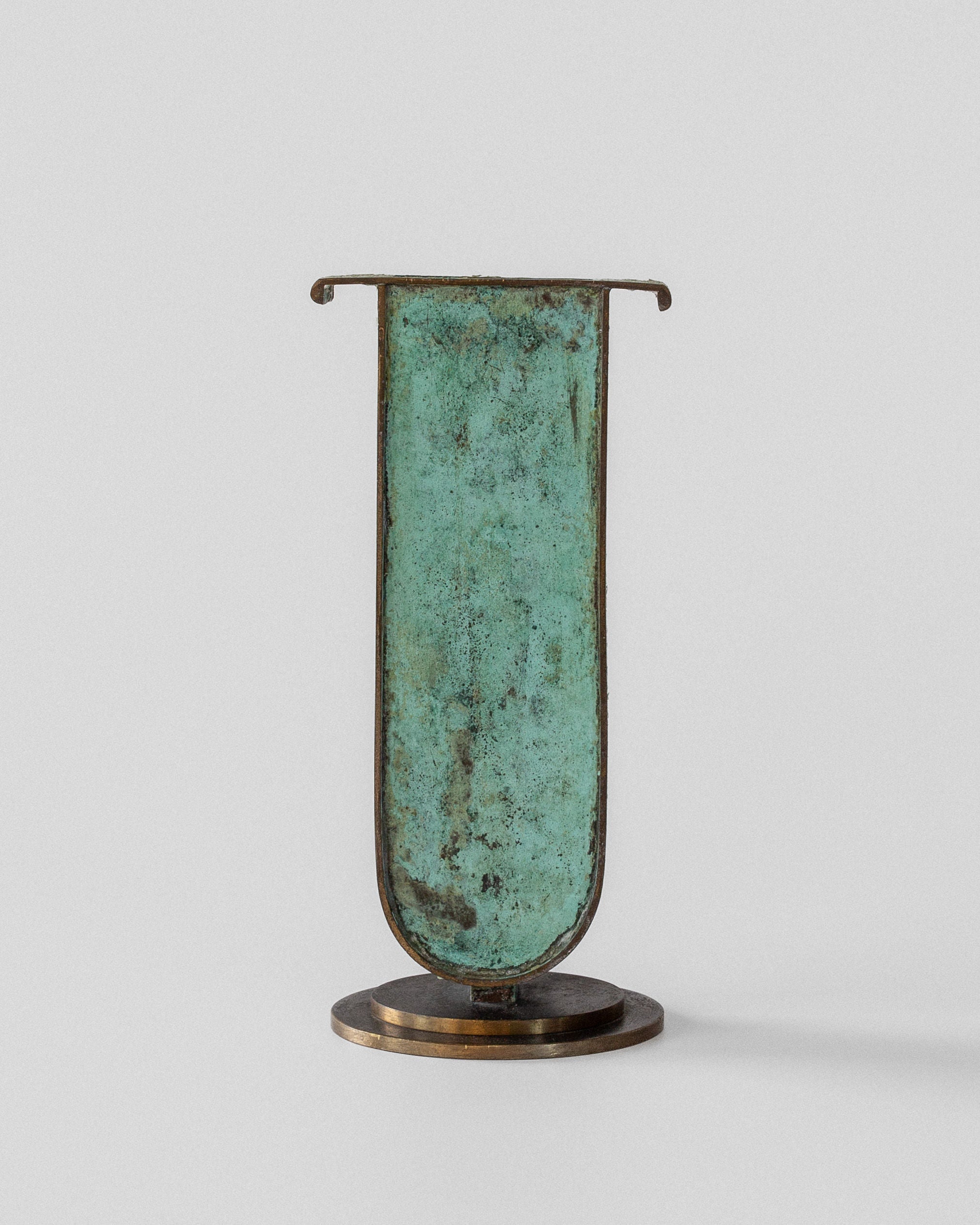 The Spigel Art Deco Brass Vase features a verdigris patina with an elongated, curved rectangular shape and a round base, offering a rustic and artistic appearance.