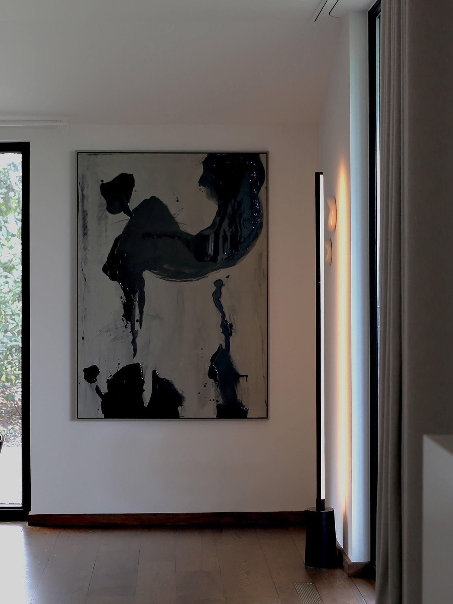 The room has a contemporary vibe, showcasing "Black Drift" by STUDIOPOLS, an abstract black and white artwork on a framed canvas. A tall, slim floor lamp stands elegantly on the wooden floor. Sunlight streams through a large window, casting delicate shadows and complementing the minimalist décor in neutral tones.