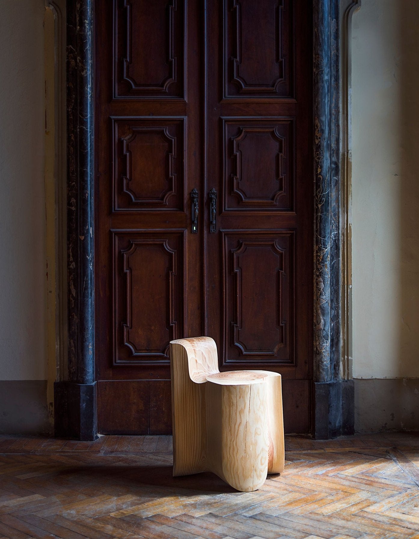 The "Chair from Wood after Storm" by Touch With Eyes rests on a decorative hardwood floor in front of an ornate, dark wooden door. The door is adorned with intricate paneling and marble trim, set beneath a softly lit, arching wall design.