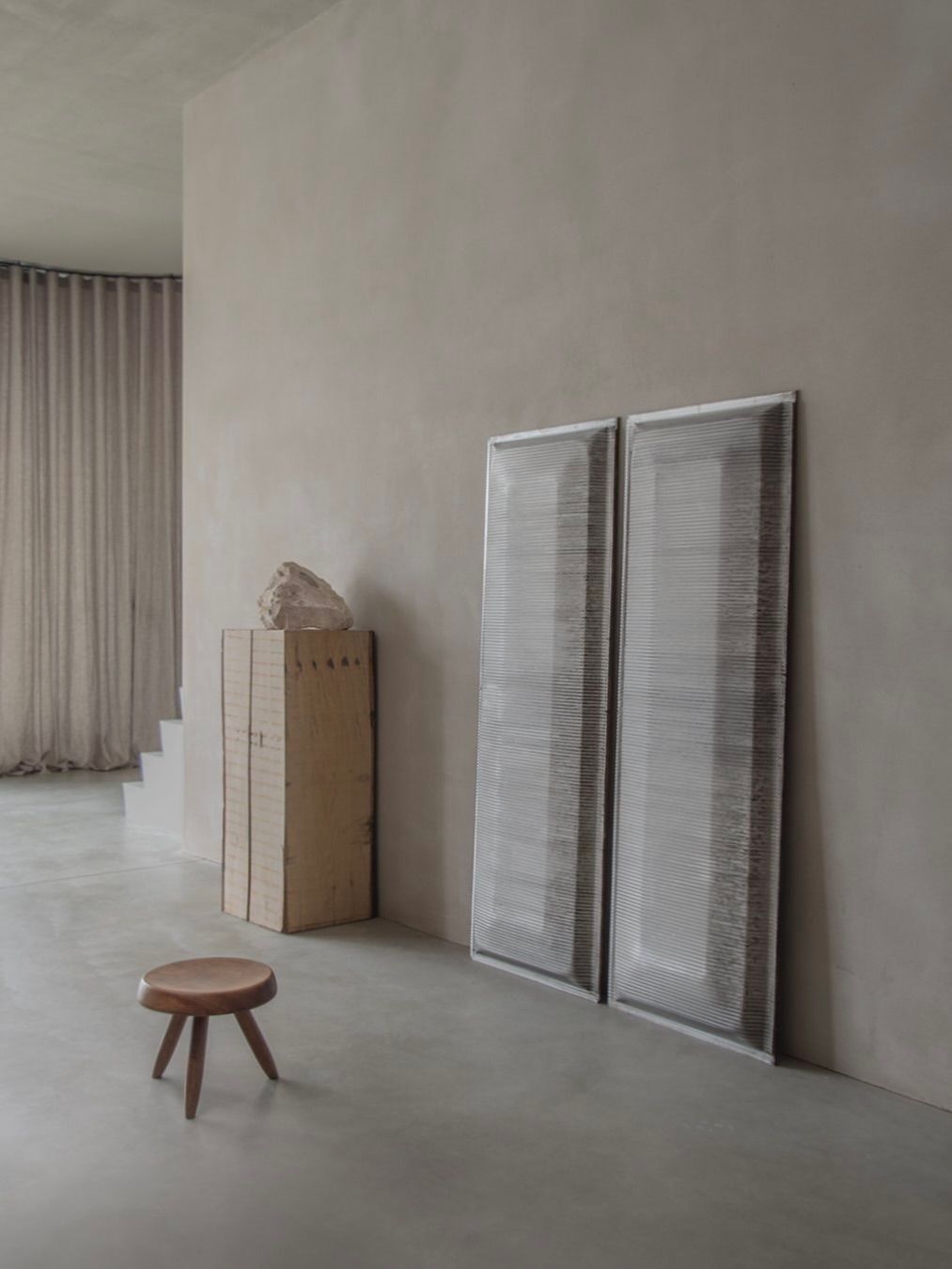 In a minimalist interior, a wooden stool rests on a smooth floor. Two tall stackable chairs evoke the style of Bicci de' Medici's Jean Prouvé - Façade Panel 70 and lean against a wall beside a tall cabinet with a large rock on top. Beige curtains partially cover the wall, revealing stairs.