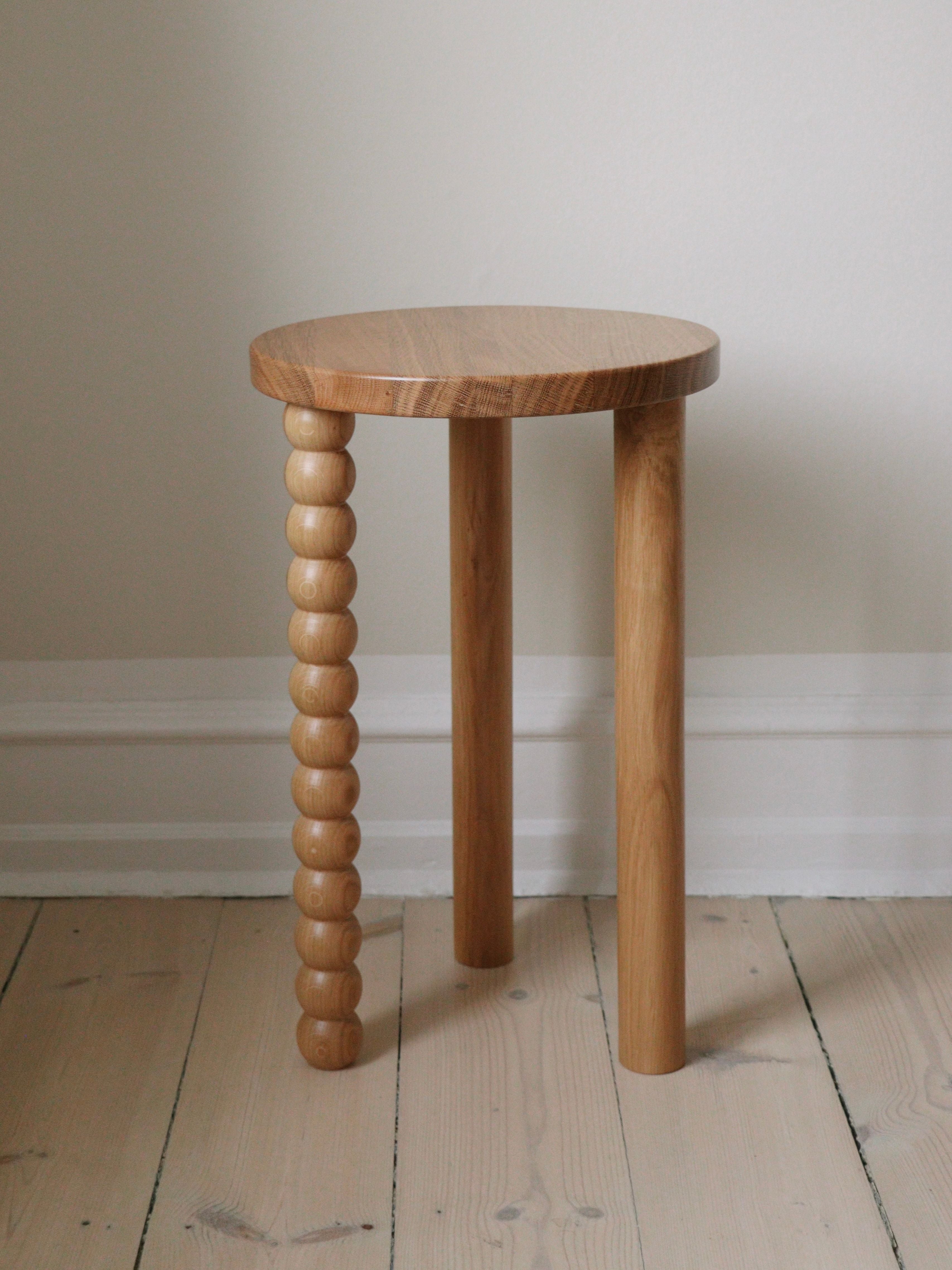 The Piloti Side Table in Oak by Maitoinen Home features a unique design with two smooth cylindrical legs and one distinctive turned wood leg adorned with a beaded pattern, placed against a light-colored wall and wooden floor.