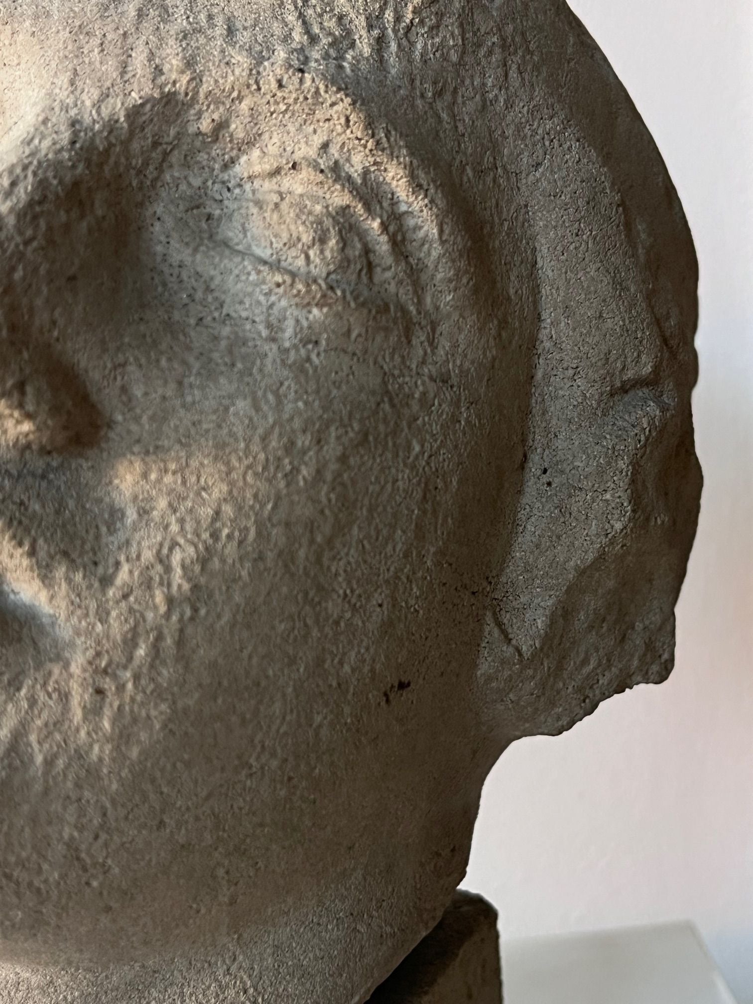 A close-up of the Médecine Decorative Bust reveals a textured sandstone composite illustrating a partial human face, highlighting an eye and nose. Its surface exhibits a rough, weathered appearance, set against a plain, light-colored wall.