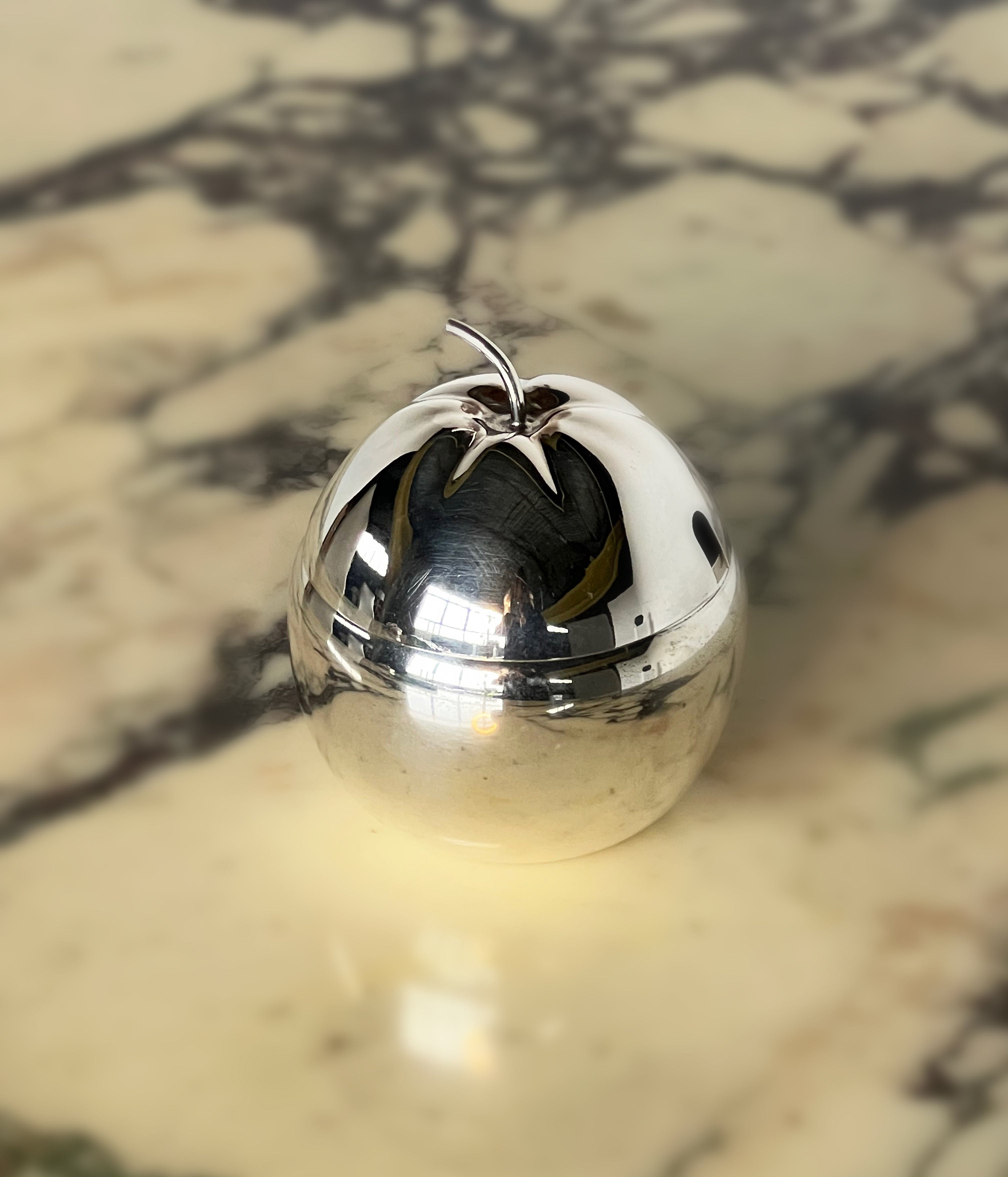 The Silver Plated Apple Jam Pot by Porter Studio, with a reflective exterior and small stem on top, rests on a marbled surface. Its shiny facade captures the surroundings beautifully, creating an intricate play of light and shadow.