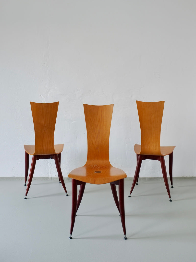Three Veter Vintage chairs, with sleek curved backs and angled legs, are set in a white-walled room with a gray floor. These 1980s Italian plywood chairs have burgundy legs and a warm finish, exuding minimalist elegance reminiscent of Philippe Starck's style.