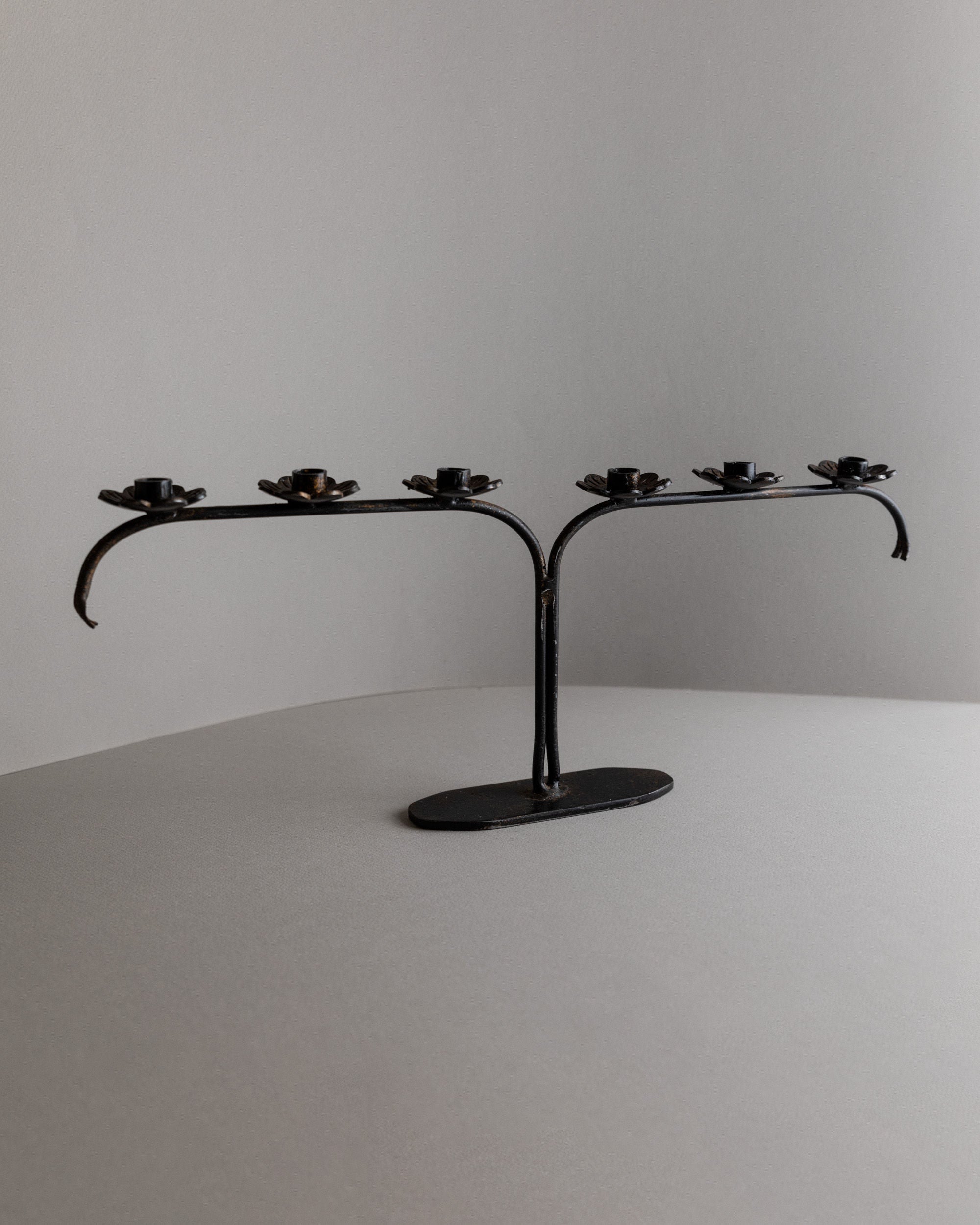The Bottega Jacobs Iron Candleholder from Italy's 1960s, featuring five circular holders on a curved base, is displayed elegantly against a plain gray background.