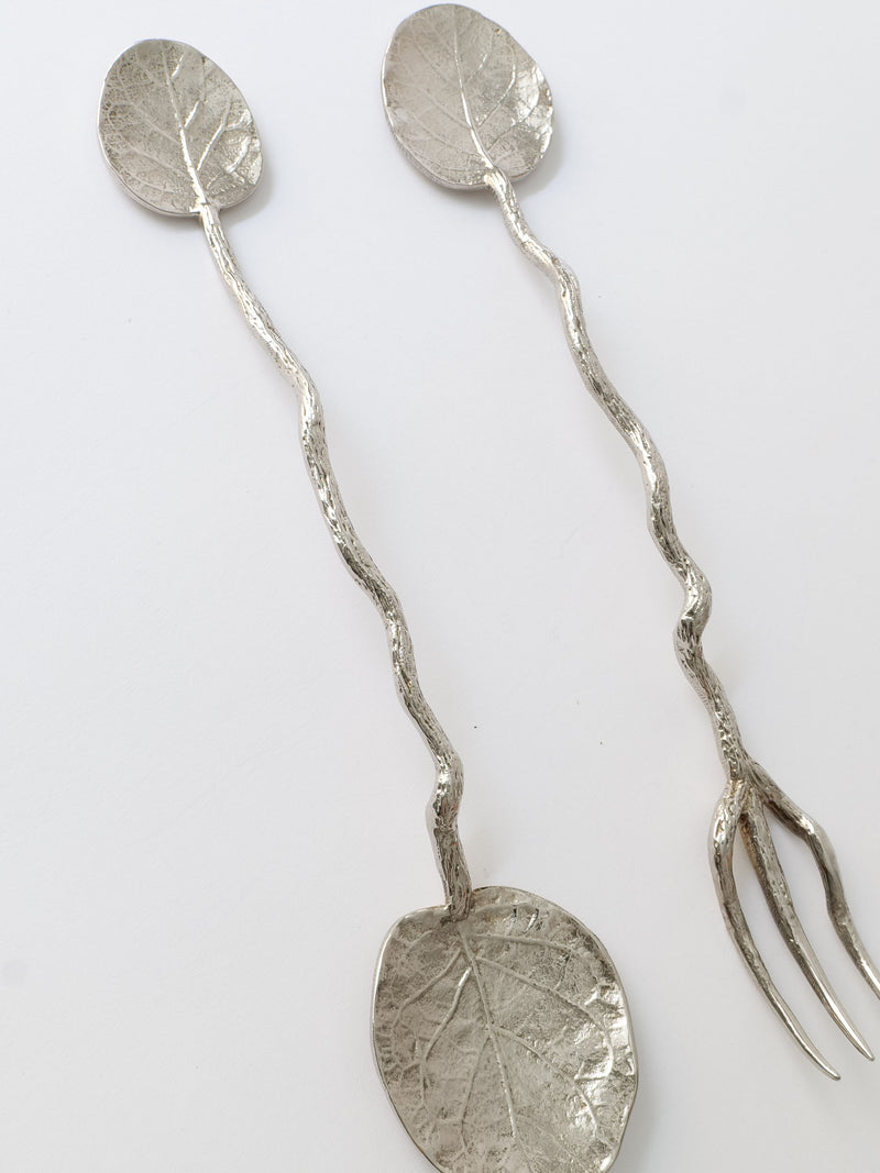Two Aram serving utensils with leaf designs and a twisted stem, one with a spoon end and the other with a fork end, against a white background by Collection apart.