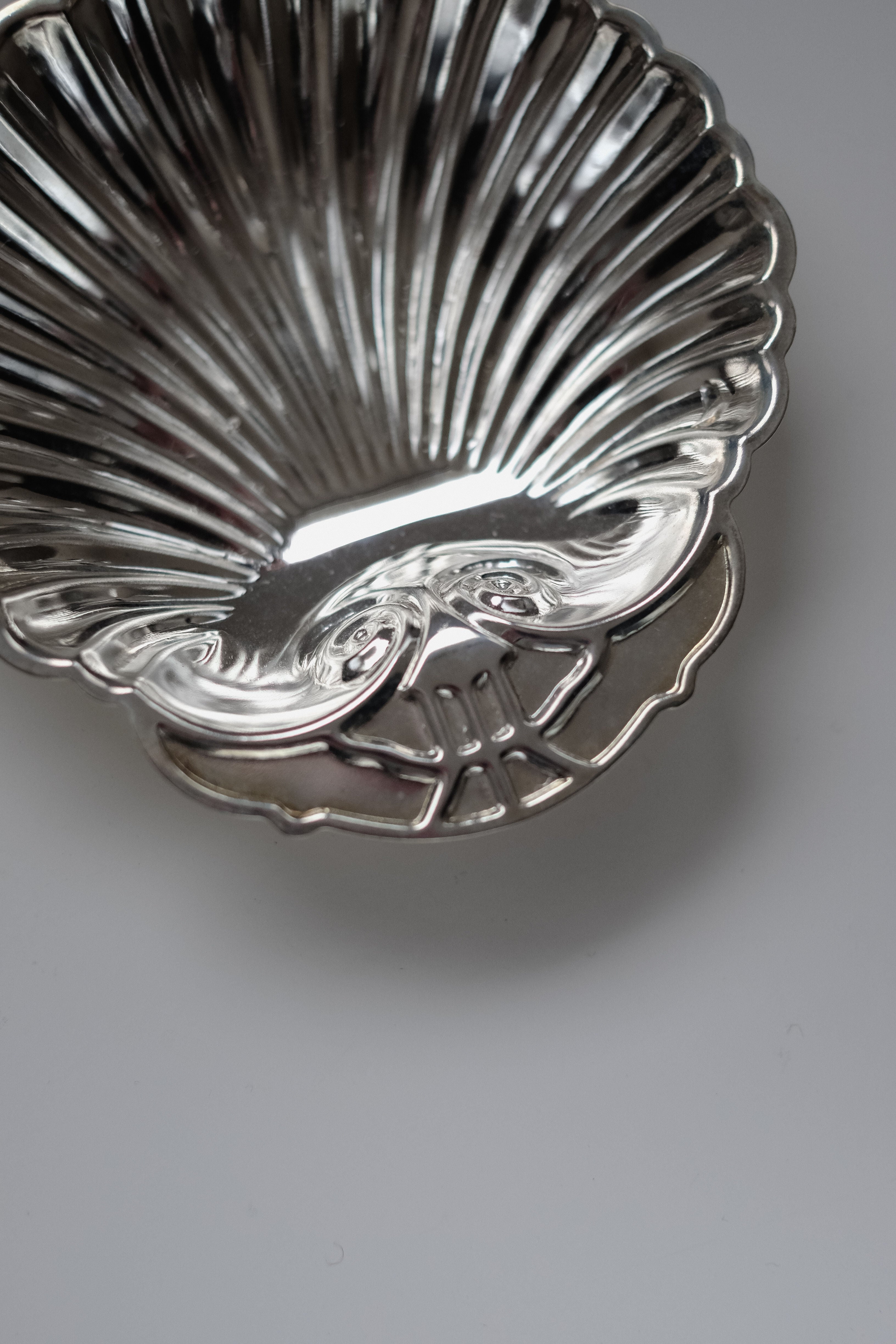 A close-up showcases the Set of 2 Silver-plated Shell Trays by septembre studios. These vintage-inspired, scallop-shaped trays feature intricate detailing and a handle at the base, making them ideal for elevating any dining setting. The background is a neutral, smooth surface.