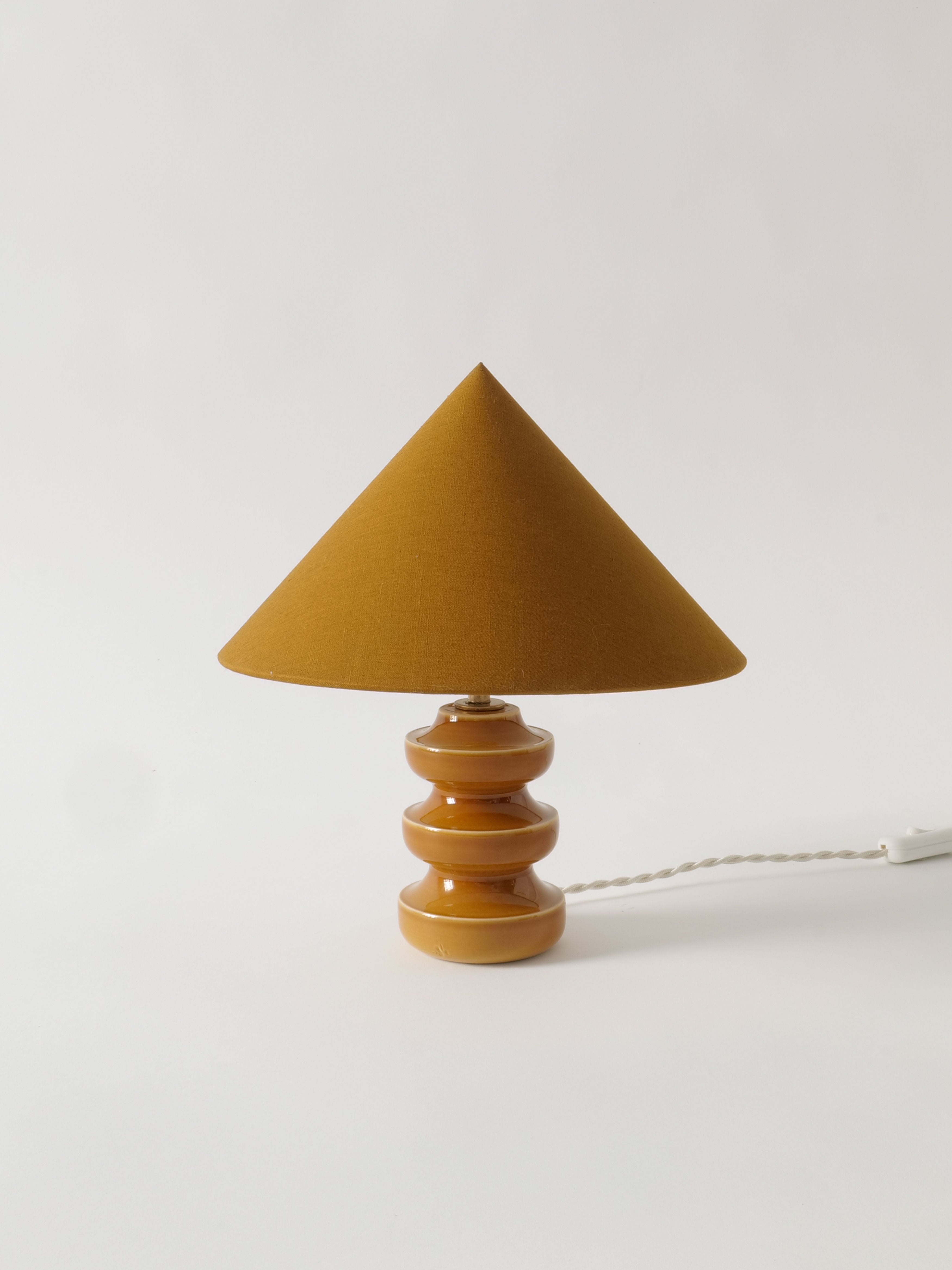 A Collection apart Small Sculptural Lamp with an earthy, conical shade and a matching ceramic base featuring three stacked circular segments. The lamp, adorned with brass fittings, is set against a plain white background with a white electrical cord extending from the base.