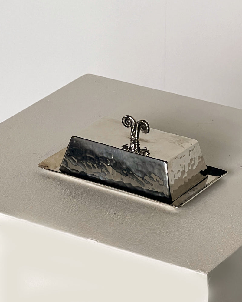 The Spiral Butter Dish by Porter Studio, a vintage 1970s piece, features a hammered silver surface with a decorative handle and rests on a square white platform, creating a textured contrast against its minimalistic background.
