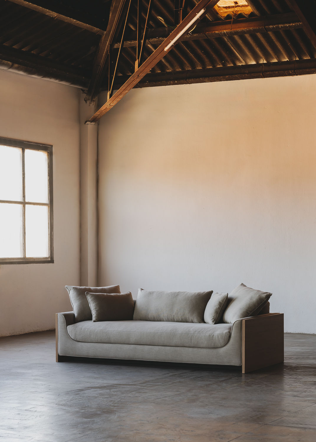 In a minimalist room with a high ceiling and an exposed beam, the ASERIES OF OBSTACLES, SL Marina Sofa features a solid oak frame that enhances natural light from a large window. With plush cushions and removable backrests, it leans against an off-white wall on a polished concrete floor.