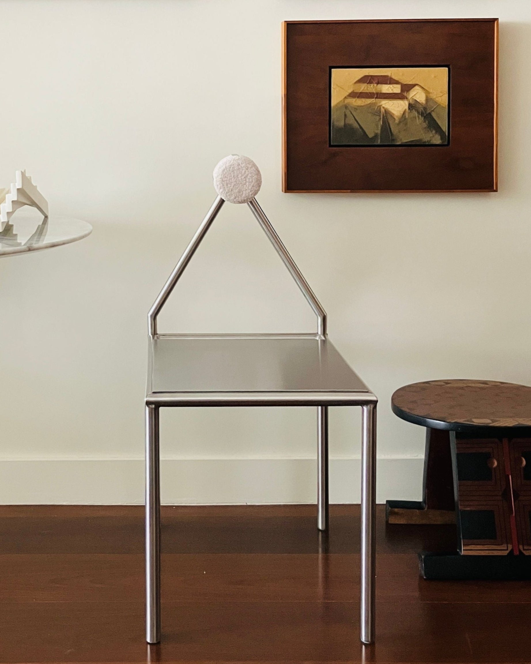 The 'H01' Stainless Steel Chair by HT, featuring a metal frame, triangular backrest, and white spherical top, sits on a wooden floor. Abstract art and geometric decor embellish the surrounding wall and surfaces.