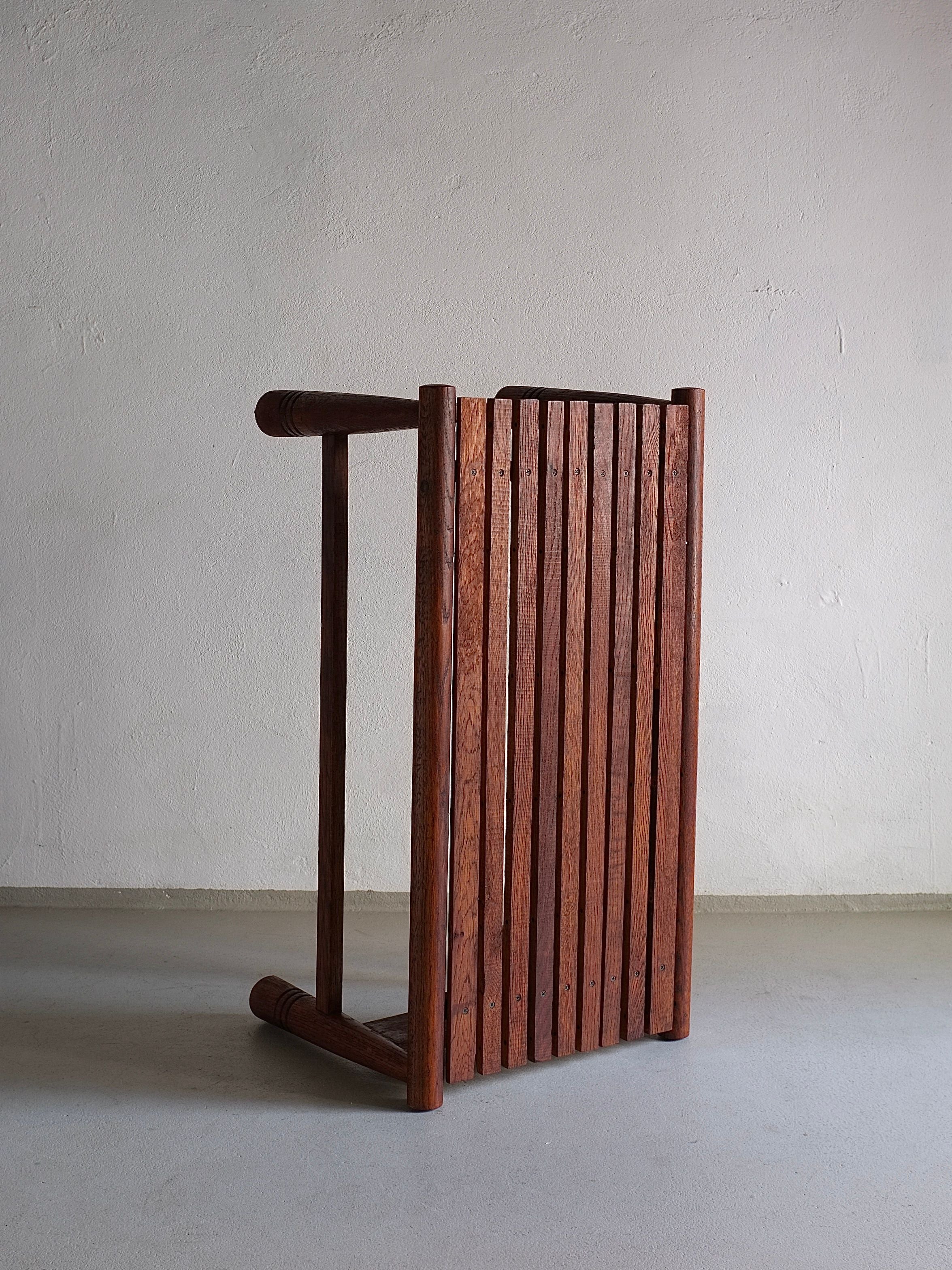 A Carved Dark Oak Bench by Veter Vintage, reminiscent of a 1960s French design, is leaning sideways against a bare, off-white wall. The bench features a slatted design with vertical slats forming the seat and cylindrical legs visible on the left side. The floor beneath is plain and light gray.