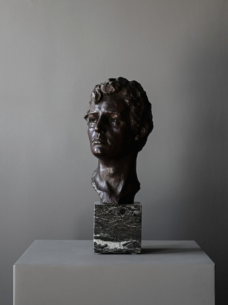 The "Bronze Bust of Male by Victor Demanet" from Studio OSKLO features a man's curly-haired head on a square marble base, displayed atop a gray pedestal against a plain gray background.