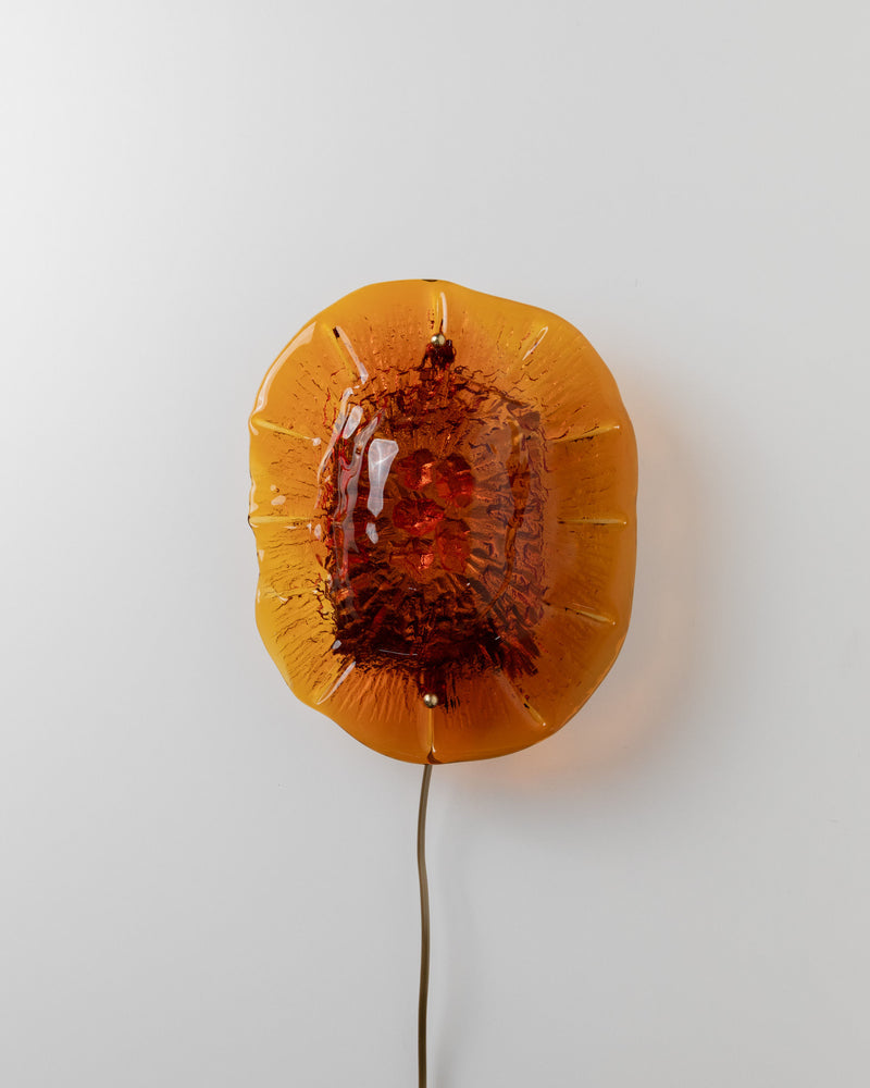A circular, amber-colored Wall Lamp by Vitrika 60s from Bottega Jacobs features a textured surface and a brown center, mounted on a white wall. A power cord extends from the bottom of the fixture, adding a nostalgic touch to any space.