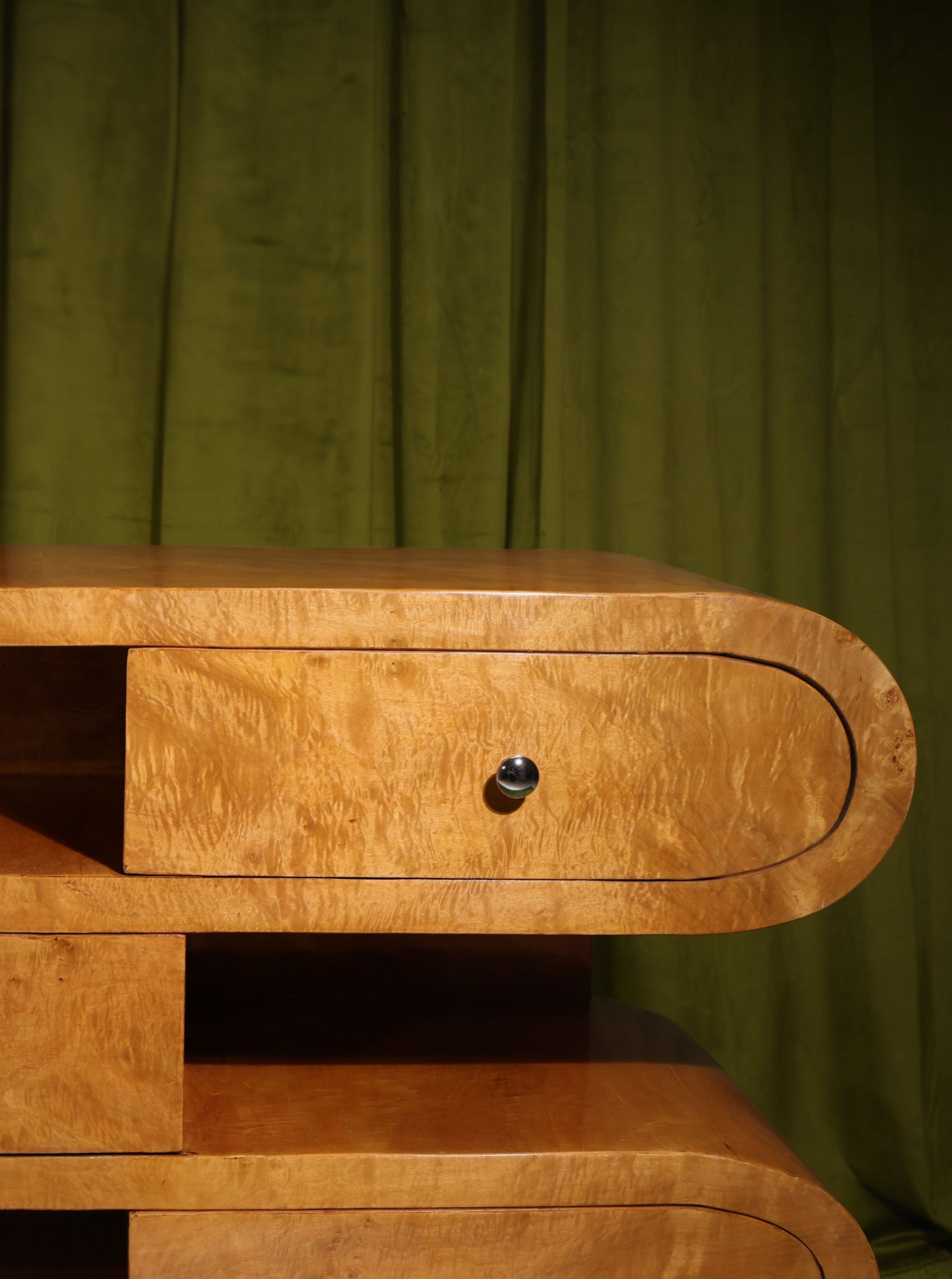 A close-up of a wooden dresser with curved edges and a sleek, polished Hebi Drawers Console Burl Wood finish from Meola Interiors. The mid-century design piece has multiple drawers, one of which is slightly open, revealing a small black knob. The background features a green curtain.