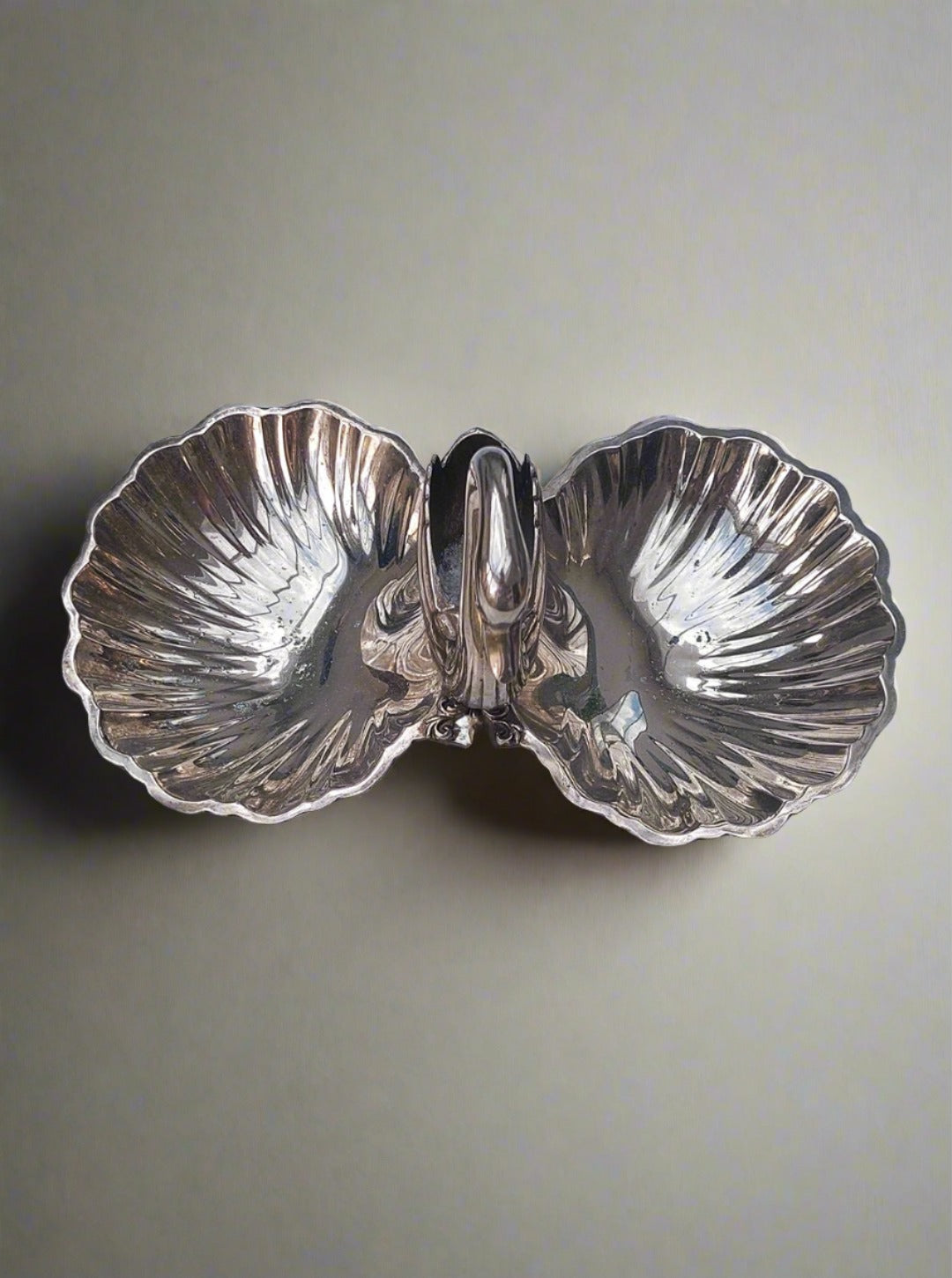 The Les Objoies Shell and Swan Dish is a silver, double-sided shell-shaped dish featuring a graceful swan figure at its center that serves as a handle, showcasing exquisite craftsmanship. This elegant dish rests on a light gray marble surface, accented by subtle oxidation marks that enhance its vintage charm.