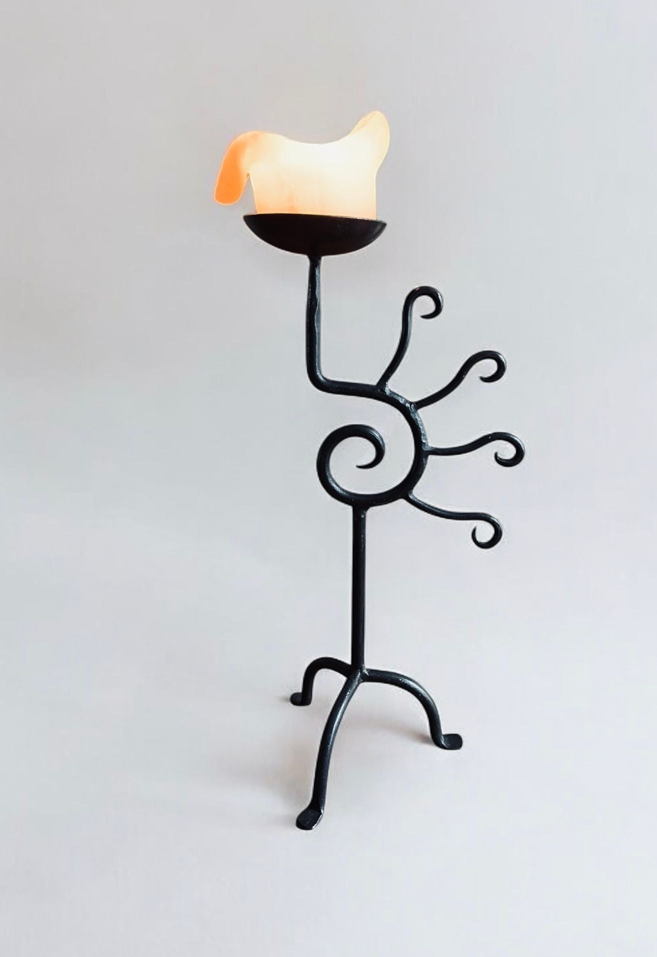 The Iron Sculptural Candleholder by Flock Studio boasts a unique spiraling black metal design with five branching arms. This modern piece elegantly displays a candle shaped like melting wax, set against a plain light gray background.