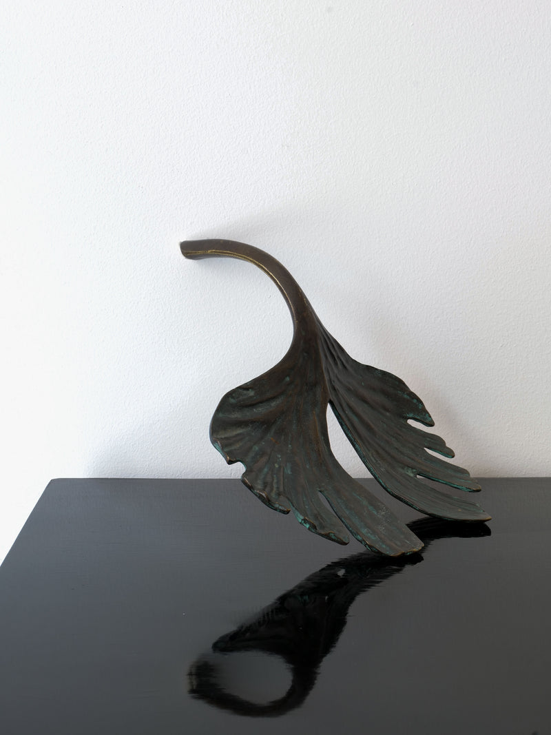 A piece from the Collection apart brand, the Bronze Jobin Sculpture, resembling abstract ginkgo biloba leaves with a smooth, dark finish, is positioned on a glossy black surface. It casts a shadow that creates a mirrored effect against both the wall and tabletop.