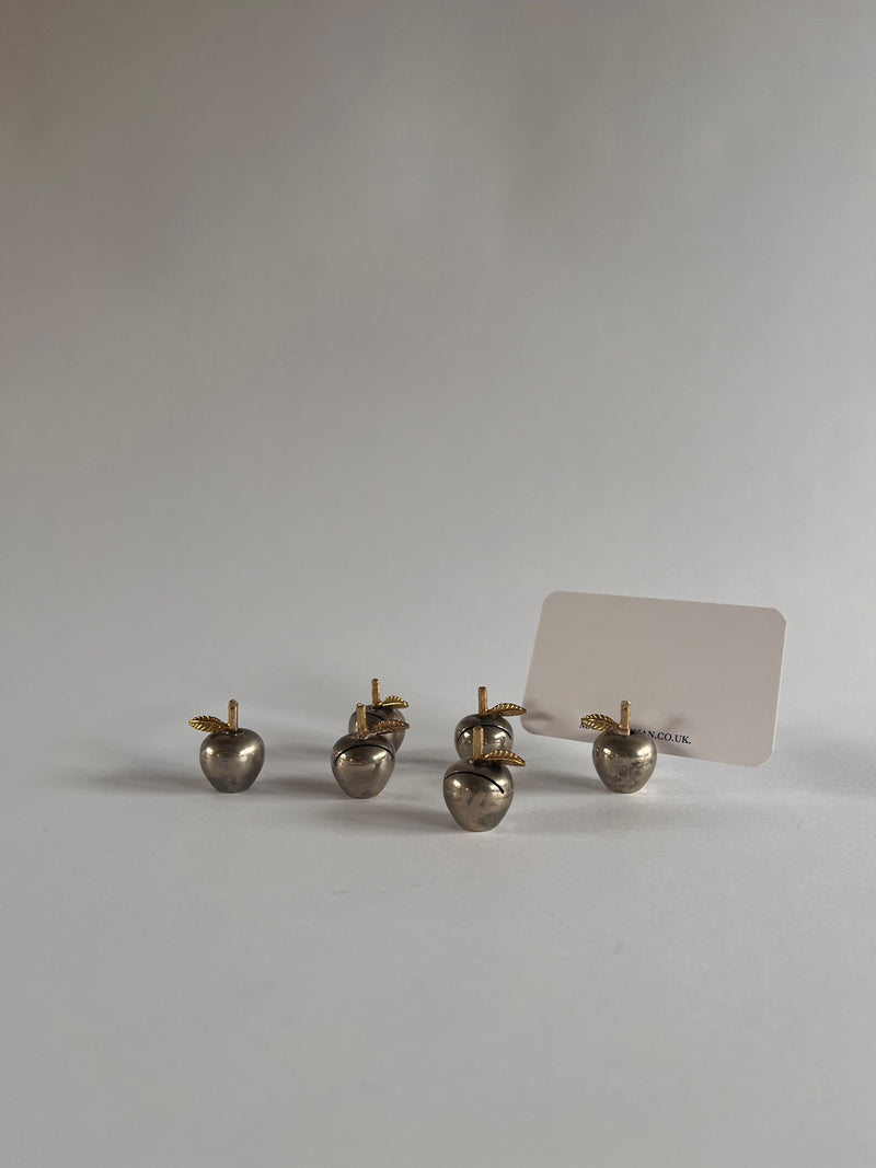 A set of six Maud Vaughan Christofle name card holders, shaped as silver-plated apples with gold stems and leaves, are arranged on a white surface. The back right apple elegantly displays a name card, with a neutral gray background enhancing the sophisticated display.