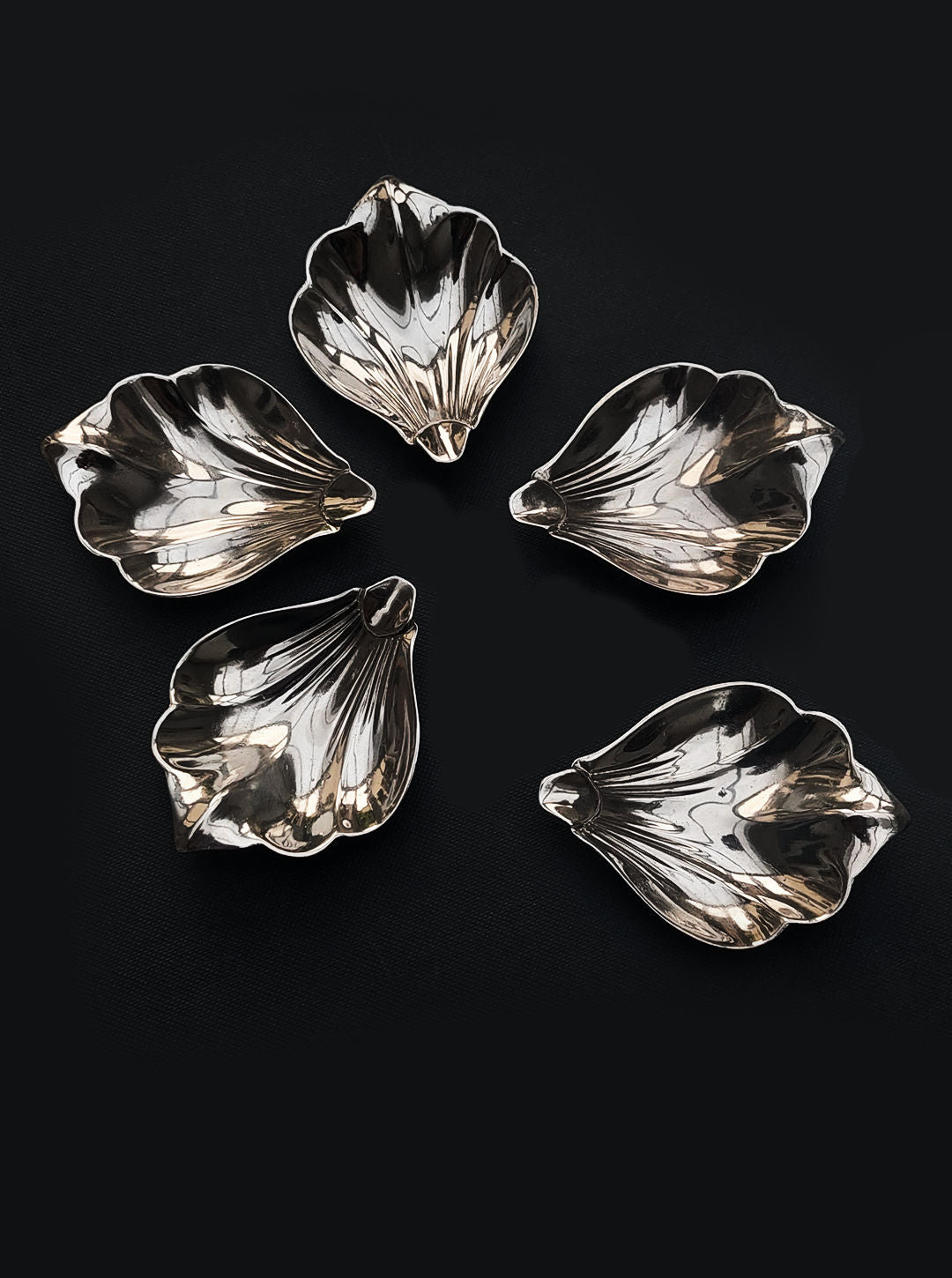 A set of five small petal dishes by Les Objoies is elegantly arranged in a circular pattern on a dark surface. Their silver, leaf-shaped design and reflective surfaces accentuate the detailed craftsmanship, making them an exquisite addition to any setting.