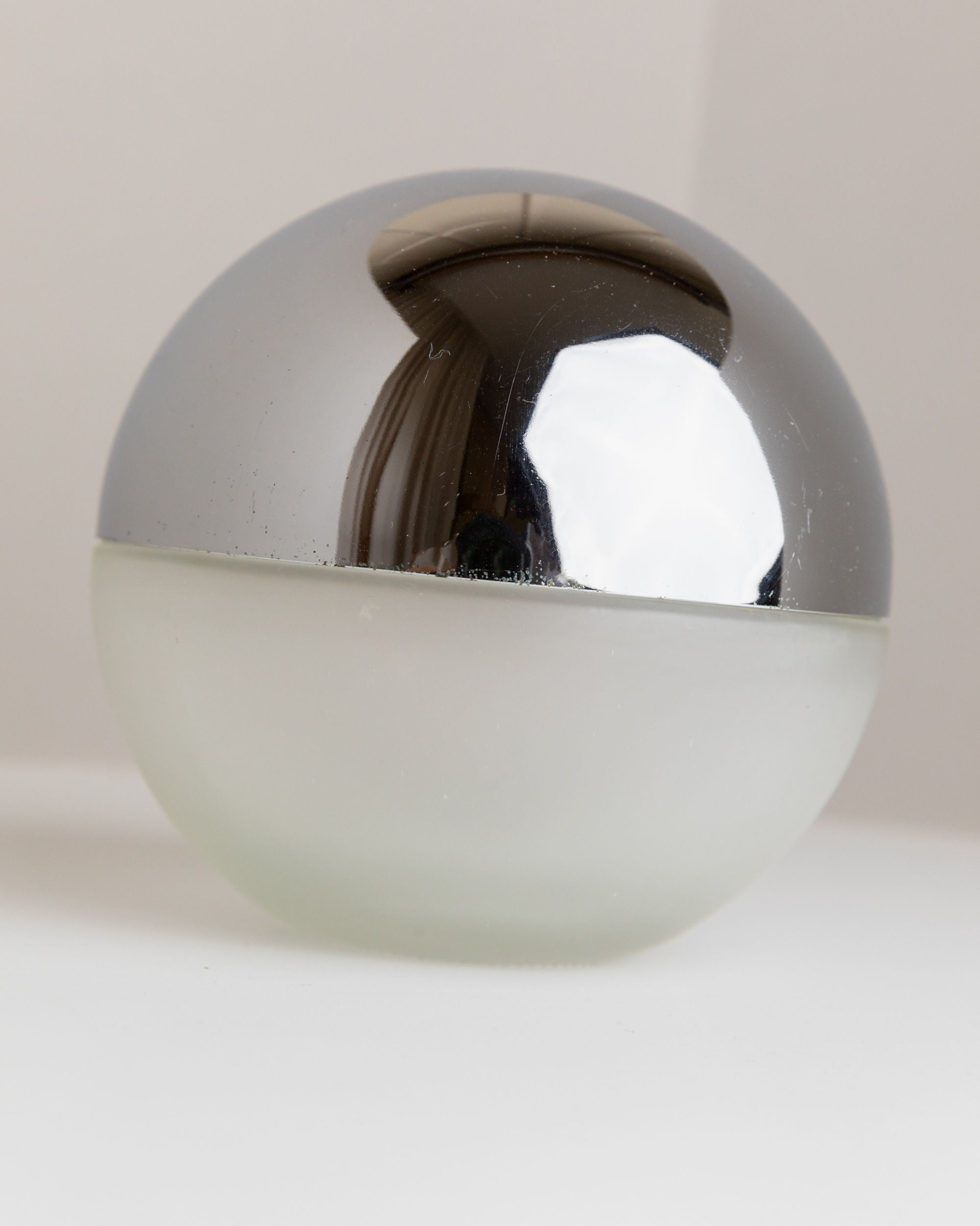 A small, spherical Bottega Jacobs container with a frosted glass lower half and a shiny metallic upper half, resembling the vintage Liqueur Bottle from Italy 1980s, sits on a plain surface.
