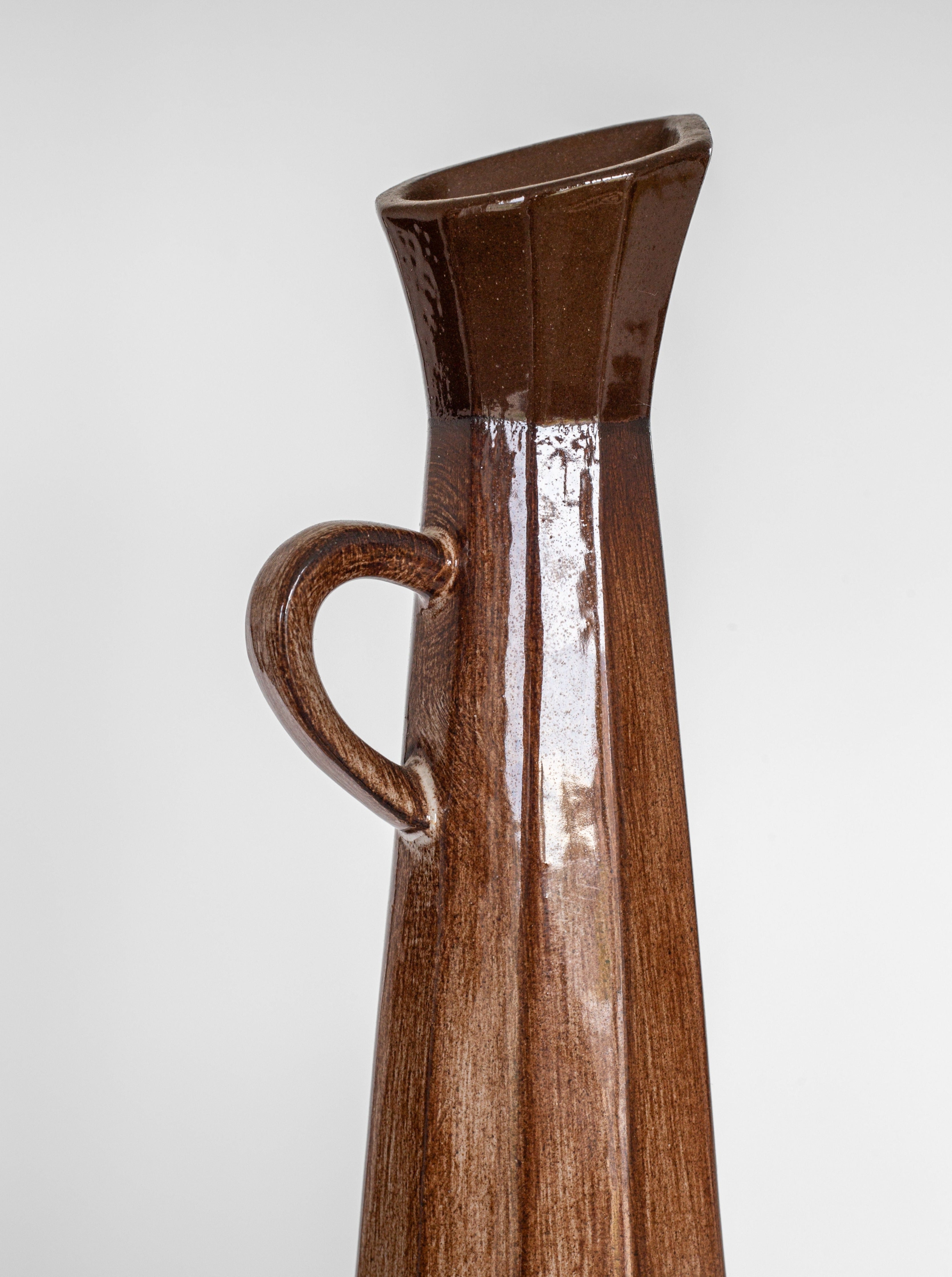 A Cubist Ceramic Pitcher by Spigel, boasting a brown glossy finish, is showcased against a plain white background. Reflecting mid-20th-century design, the pitcher features a narrow, slightly slanted opening and a small handle on one side, giving it an asymmetrical and contemporary appearance.