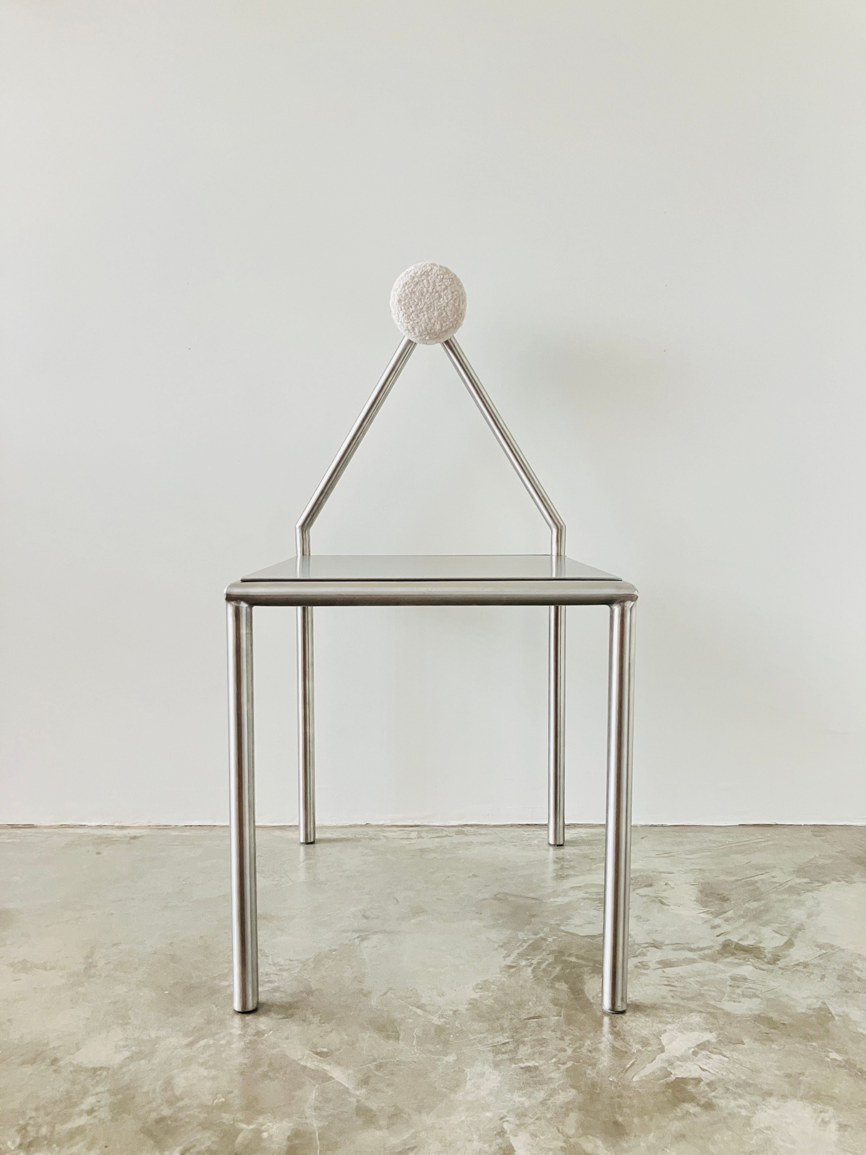 The 'H01' Stainless Steel Chair by HT features four cylindrical legs, a flat square seat, and a triangular backrest with a removable cushion, shown against a plain white wall and concrete floor.