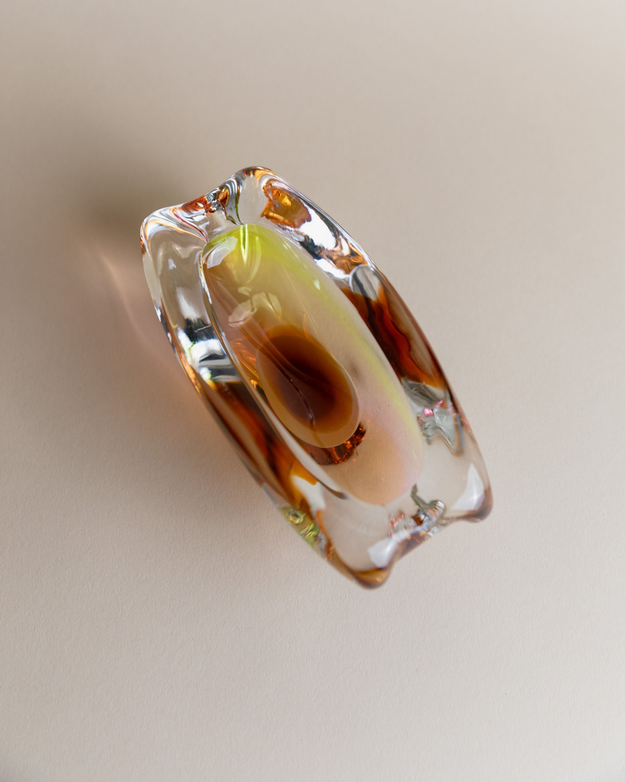 The Glass ashtray by Josef Rozinek for Novy Bor, branded by Bottega Jacobs, features a smooth, abstract design with a rounded hollow center. Its clear and amber tones evoke retro glassware's warmth and reflectivity, beautifully showcased on a light, neutral background.