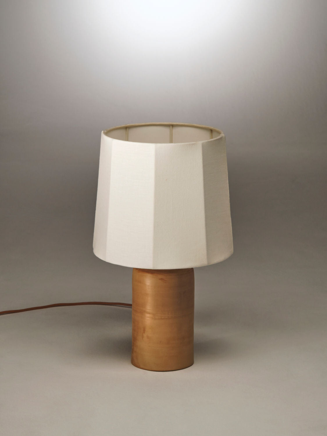 The Sonora Linen Table Lamp by Nassi Lamps with a terracotta base and a white, faceted lampshade. The lamp's electrical cord is visible, extending from the base toward the left. The background is a plain, light gray surface with a gradient, giving it a subtle spotlight effect reminiscent of Puglia's serene charm.