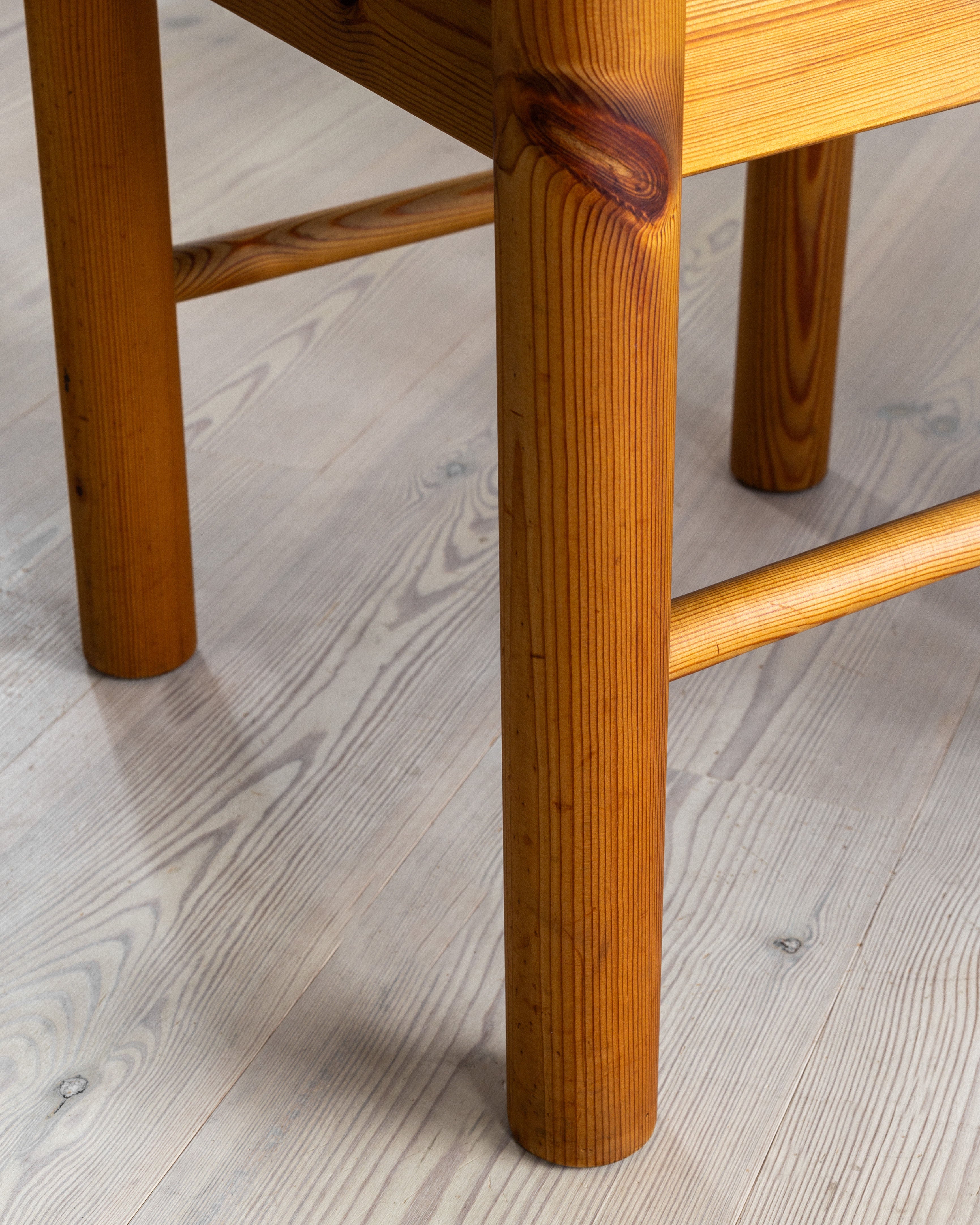 Close-up of a wooden chair leg. The chair, crafted by Spigel, is part of the "4 Pine Chairs by Rainer Daumiller" collection from Denmark in the 1970s and features solid pine wood with a natural finish that showcases its visible wood grain. The leg stands on a light wooden floor, rich with distinct patterns and grains, echoing the craftsmanship seen in these iconic Rainer Daumiller chairs.