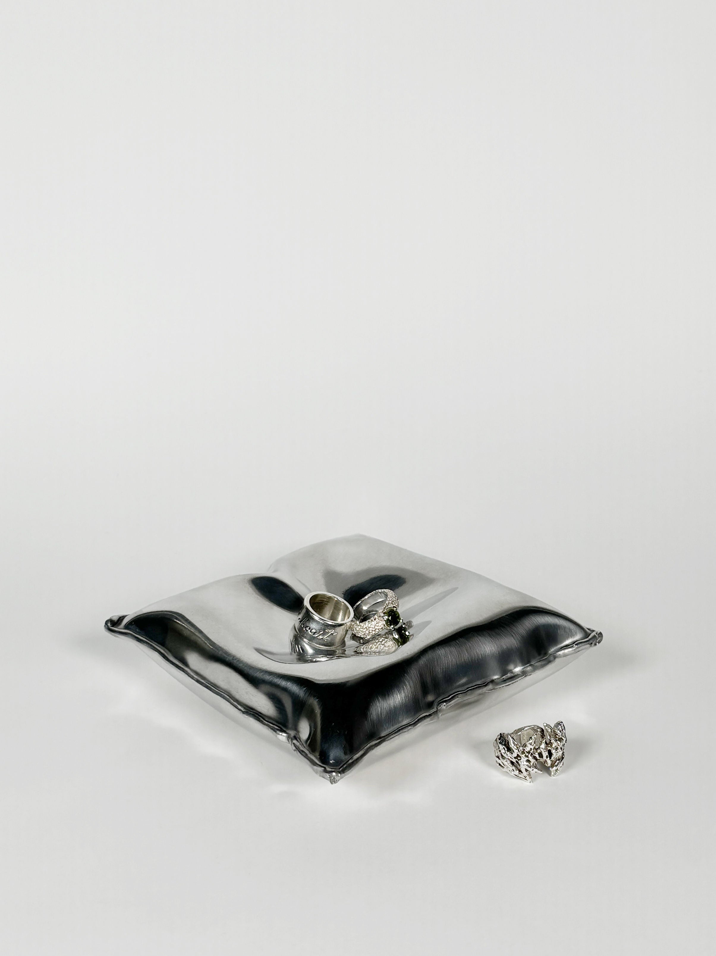 An Emma Stocklassa Pillow Bowl Large, crafted in silver aluminum, elegantly holds two intricately designed rings. Another handcrafted ring from Stockholm complements the display on a plain white background, creating a minimalist and elegant scene.