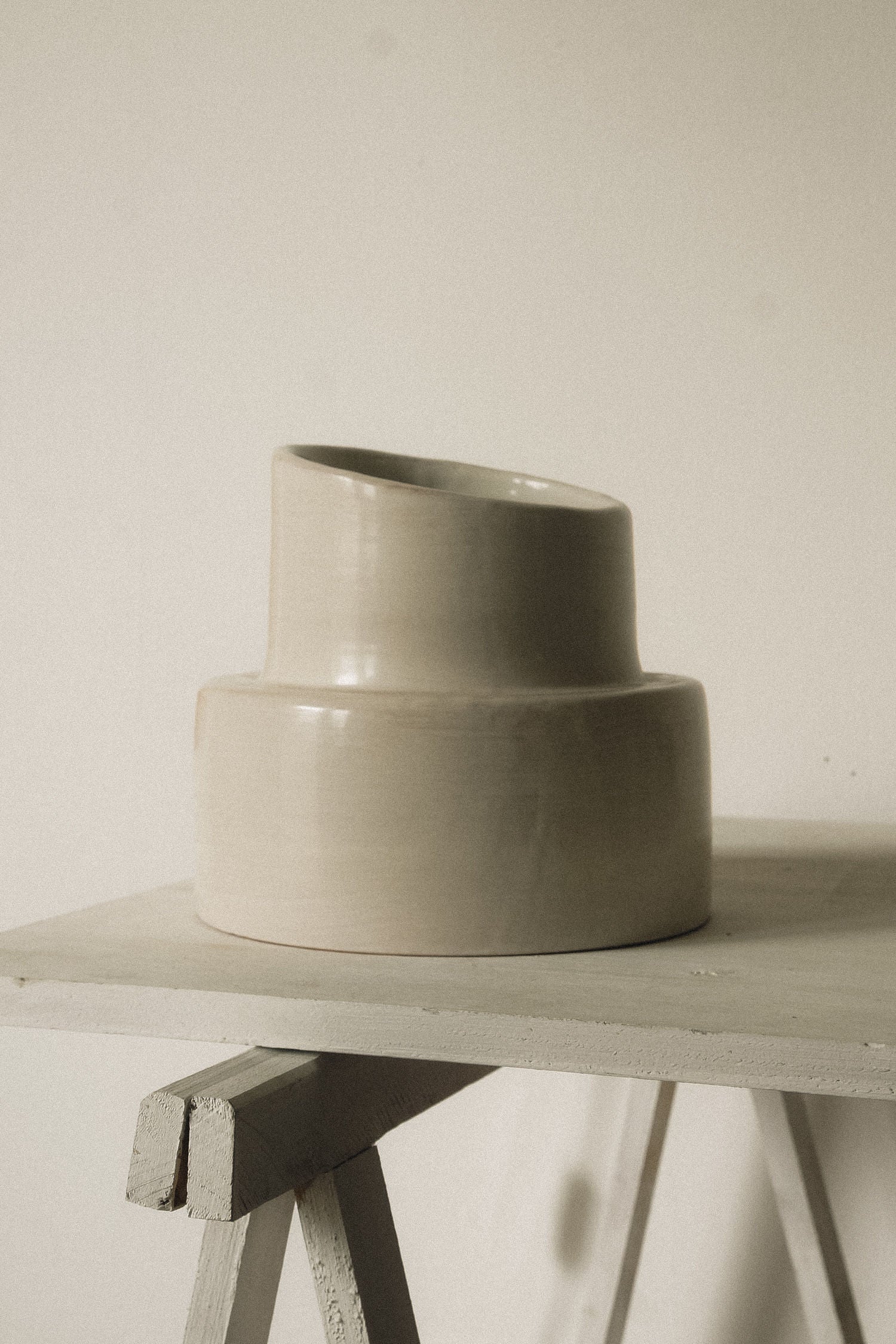 A beige modern ceramic vase with an asymmetrical neck rests on a light wooden surface next to COSEINCORSO's "Pillar" Table Lamp, set against an off-white background. Soft lighting enhances the minimalist ambiance.