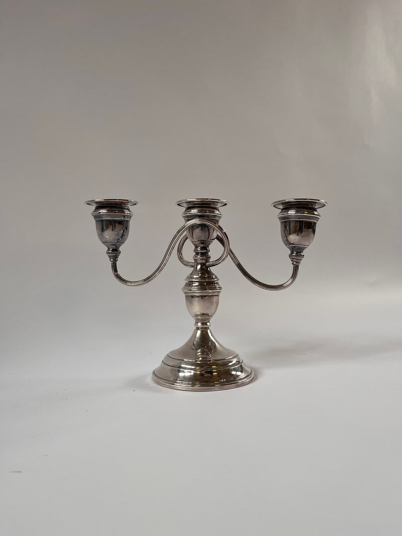 The Maud Vaughan Vintage Silver Plate Candelabra, with its shiny finish, is set against a light gray background. Featuring elegantly extended arms from the central stem, each holds a single candle holder. Its classic and elegant design exudes timeless charm.