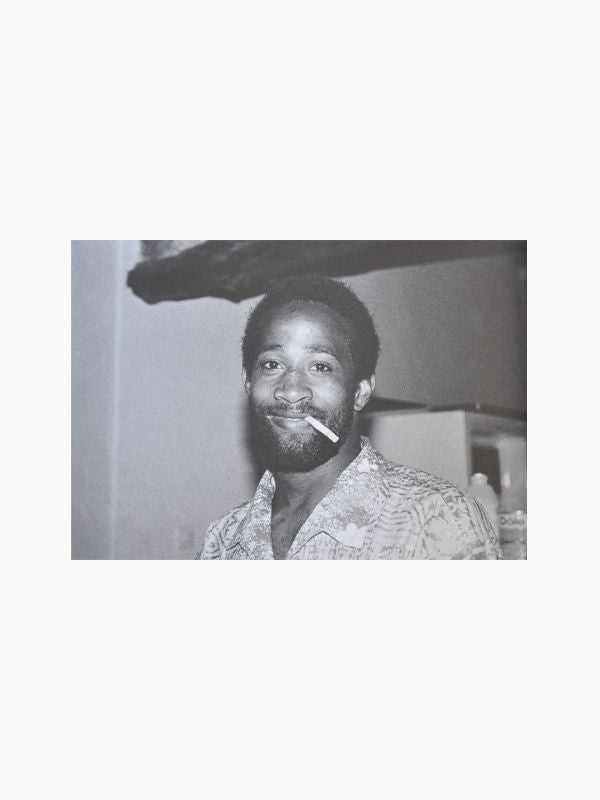Black and white image of a smiling person with a beard, wearing a patterned shirt, and holding a cigarette between their lips. The background shows a room with indistinct objects, evoking the visual poetry reminiscent of Maison Plage's "Ibiza Memories by Henry Roy.