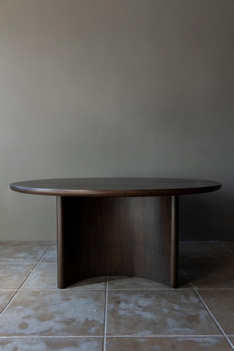 The 'Wright' Dining Table by Studio OSKLO, a minimalist handmade dark wood oval piece with a thick curved base, is placed on a tiled floor against a neutral wall.