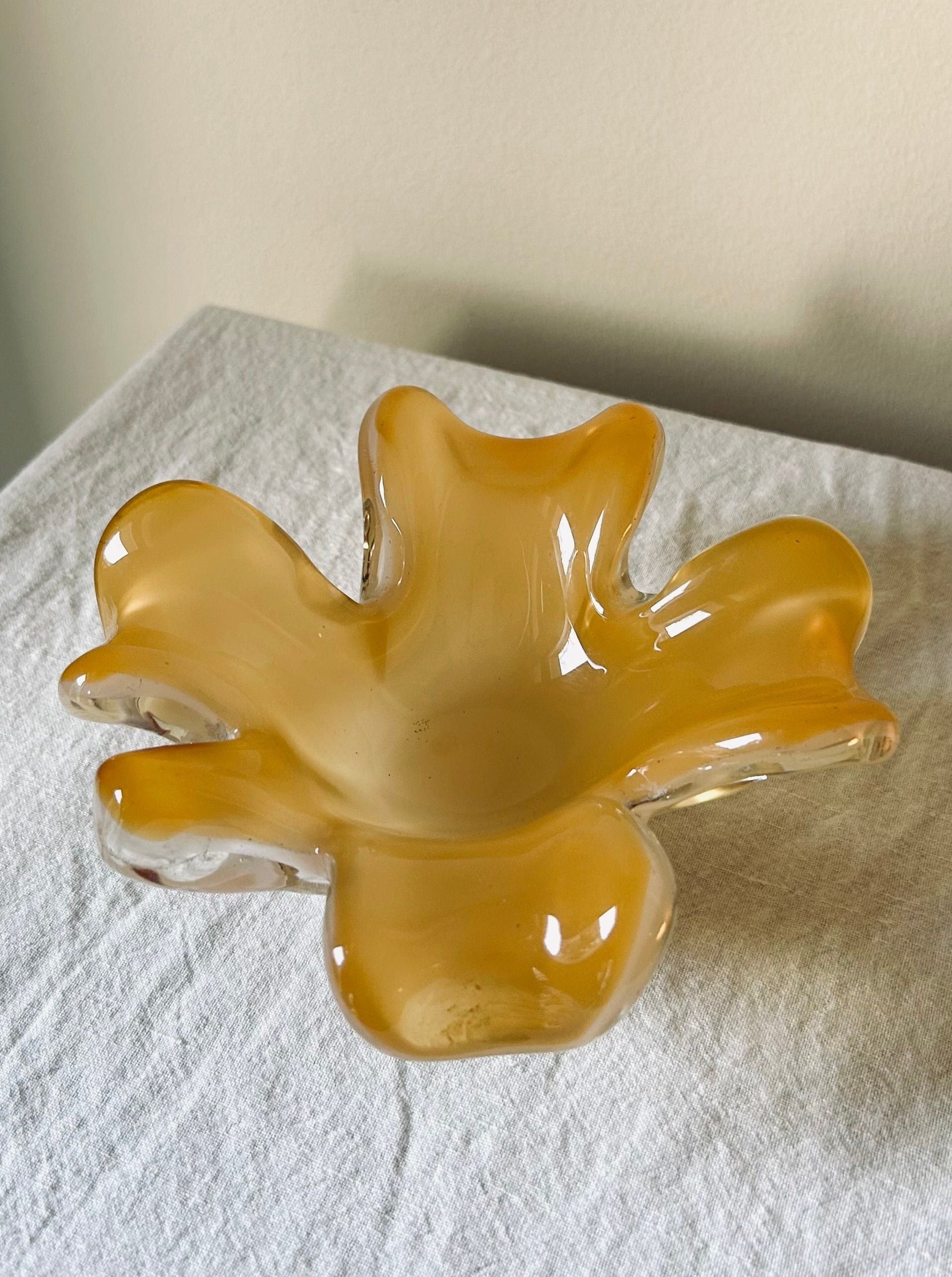 A wavy, amber-colored Murano Glass Dish from Flock Studio with a glossy finish is placed on a white textured cloth. The dish's organic, flowing shape resembles a flower with multiple curved petals, showcasing the elegance of an objet d'art created using the sommerso technique.