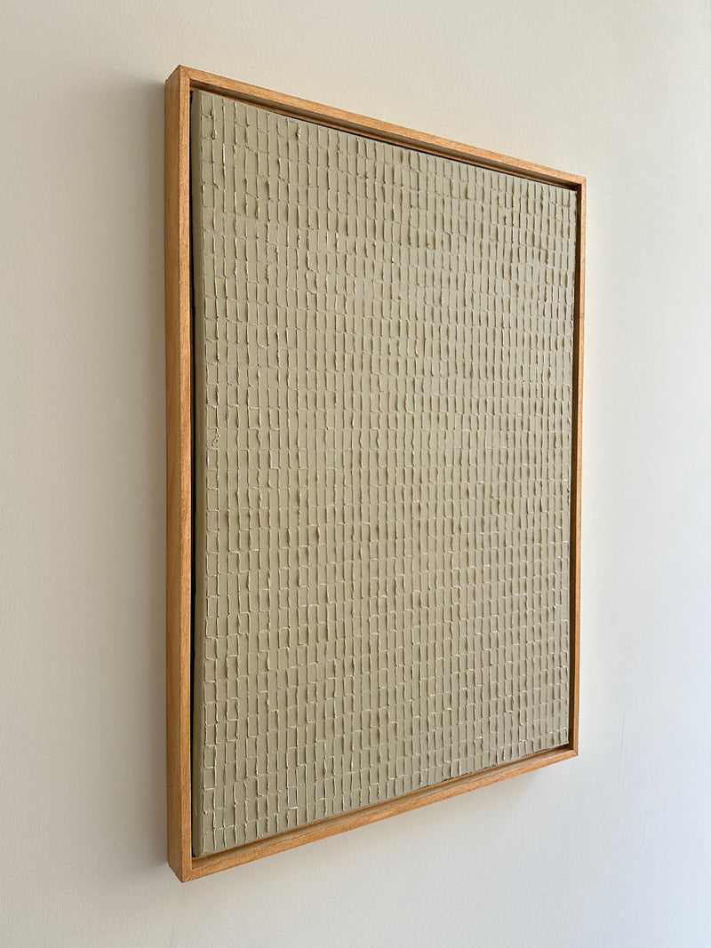 A minimalist wall art piece featuring a textured, grid-like pattern in neutral tones, framed by a simple wooden border. Inspired by Korean Dansaekhwa, Buff Titanium - Oil on canvas by Roi Elmaliah is mounted against a plain, light-colored wall, creating a clean and modern aesthetic that promotes mindfulness.
