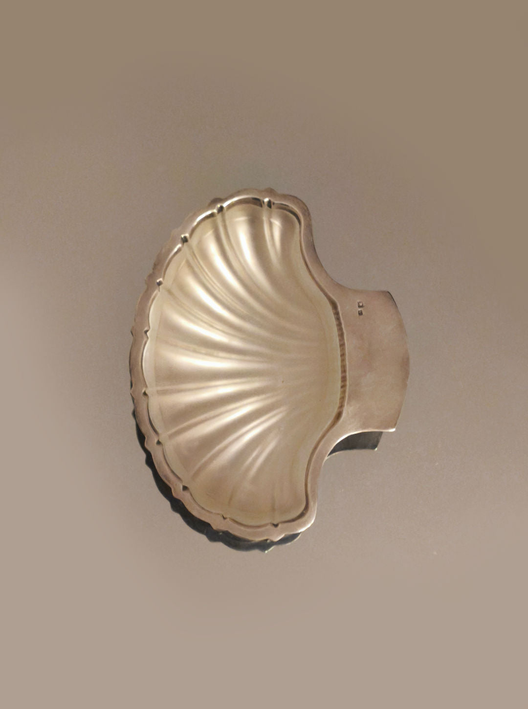 The Les Objoies Shell Butter Dish, silver-plated and shell-shaped with a wavy texture and flat handle, graces a plain background. This elegant piece adds sophistication to any table setting.