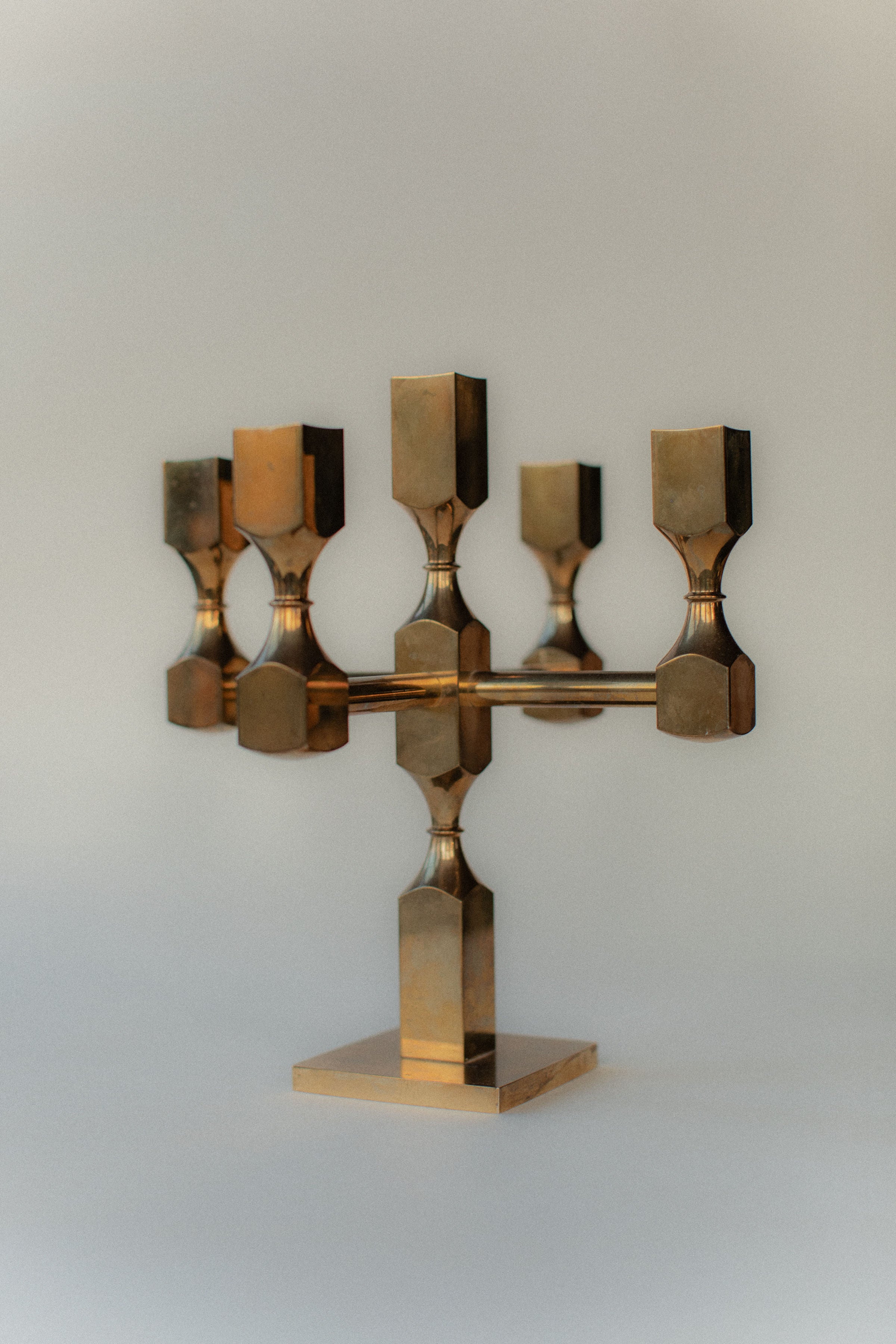 The Brutalist Brass Candleholder by Out For Lunch showcases a modern, geometric design with five candle holders on a square base, set against a light neutral background for an industrial aesthetic.